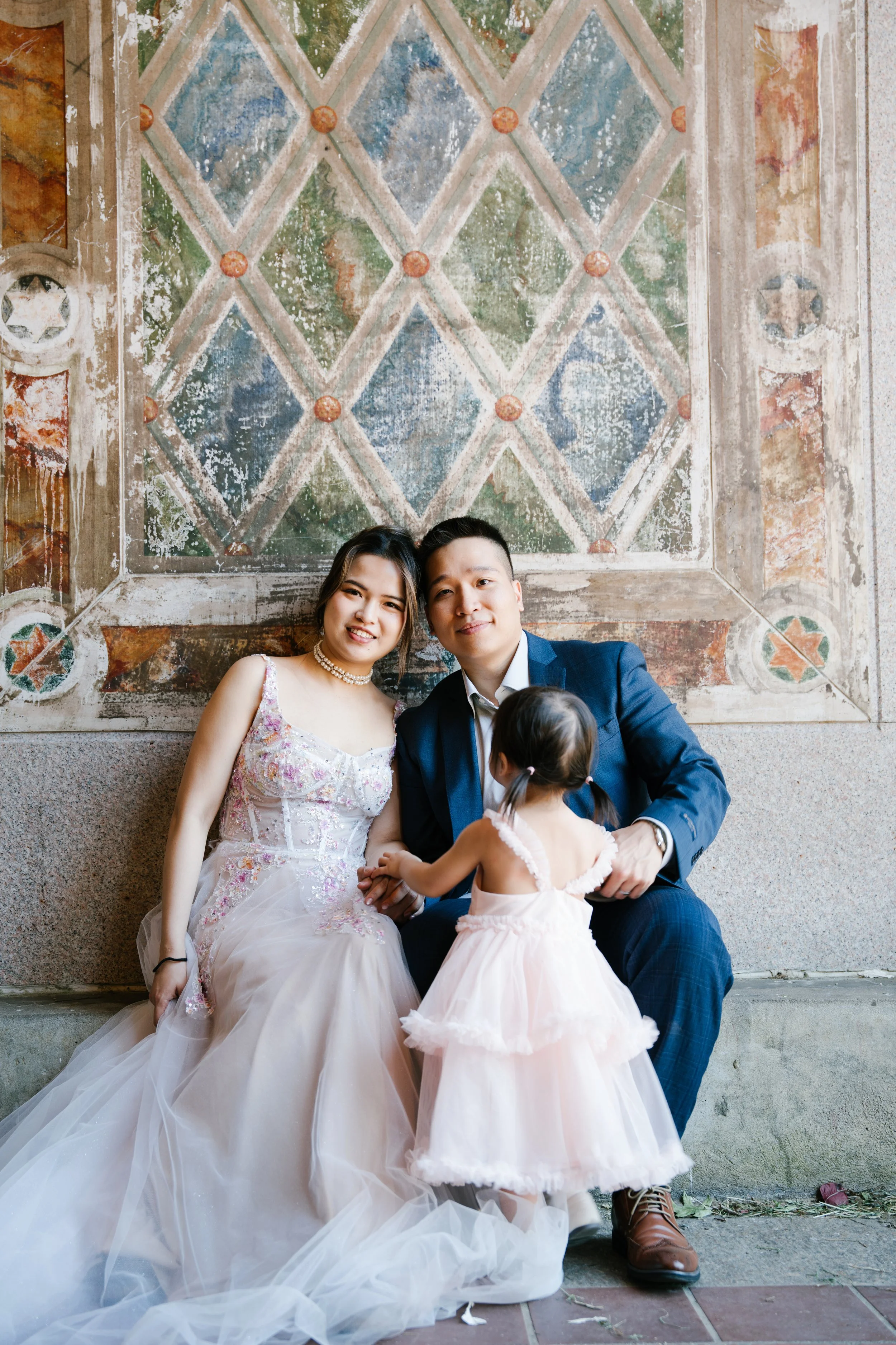 A newlywed couple with a young girl sitting against a worn fresco wall with geometric pattern and floral details. The bride wears a white floral wedding gown and pearl necklace. The groom wears a blue suit and brown shoes. The girl wears a light pink