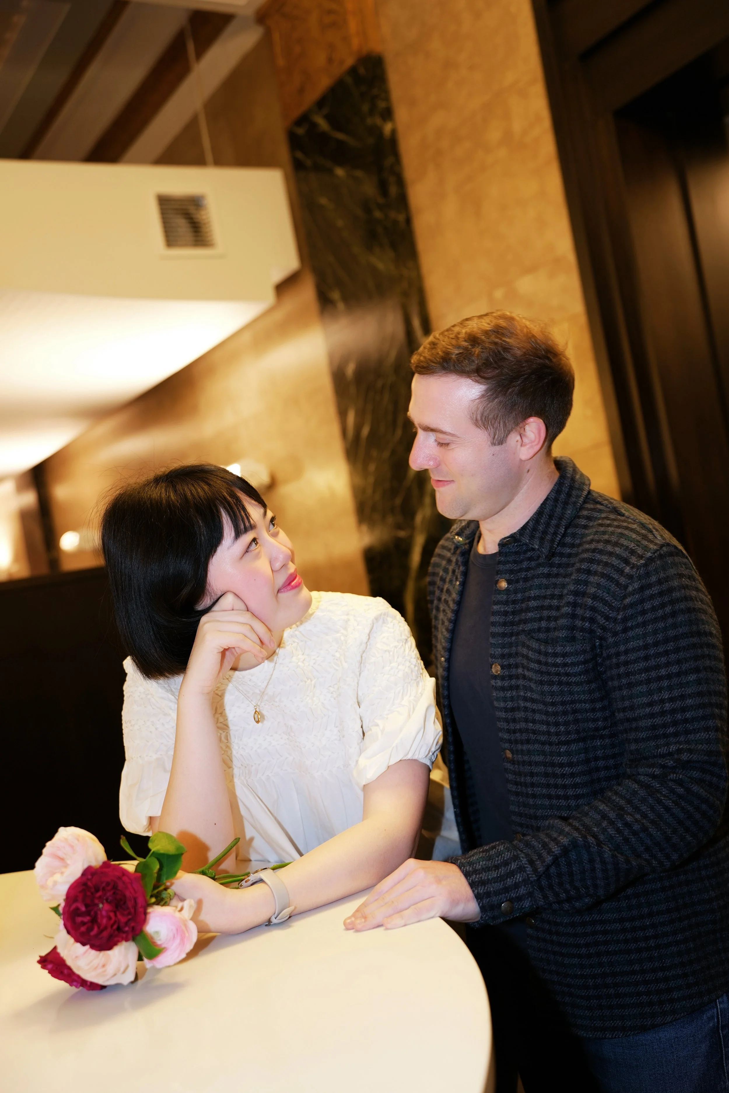 A woman with short black hair and a man with short brown hair are looking at each other, smiling affectionately, in a warmly lit room. The woman's hand rests on her cheek, and a bouquet of red, pink, and cream flowers is on the table in front of her.