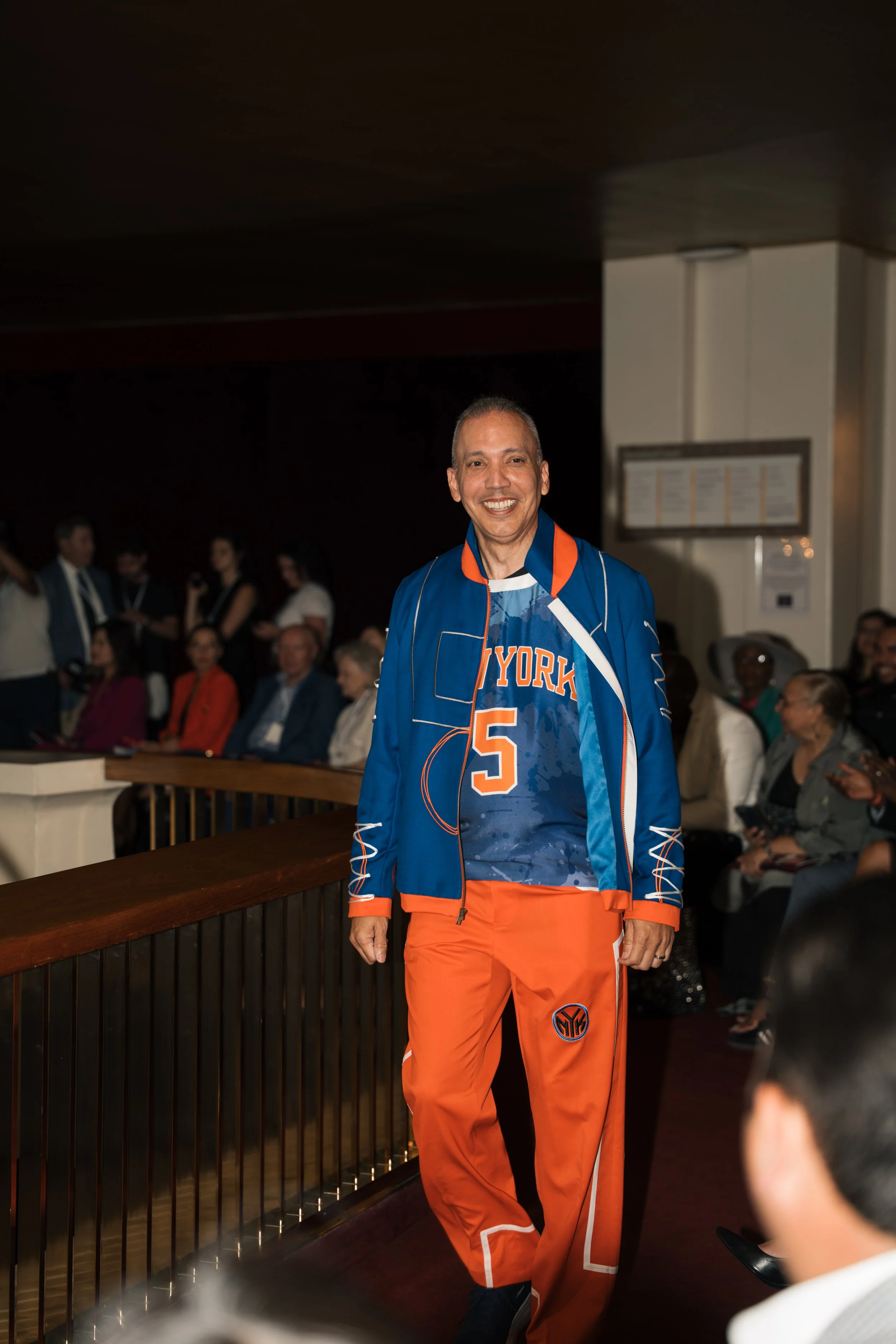A smiling man in a New York Knicks jacket and orange pants standing in a crowded room, with people seated and standing around.