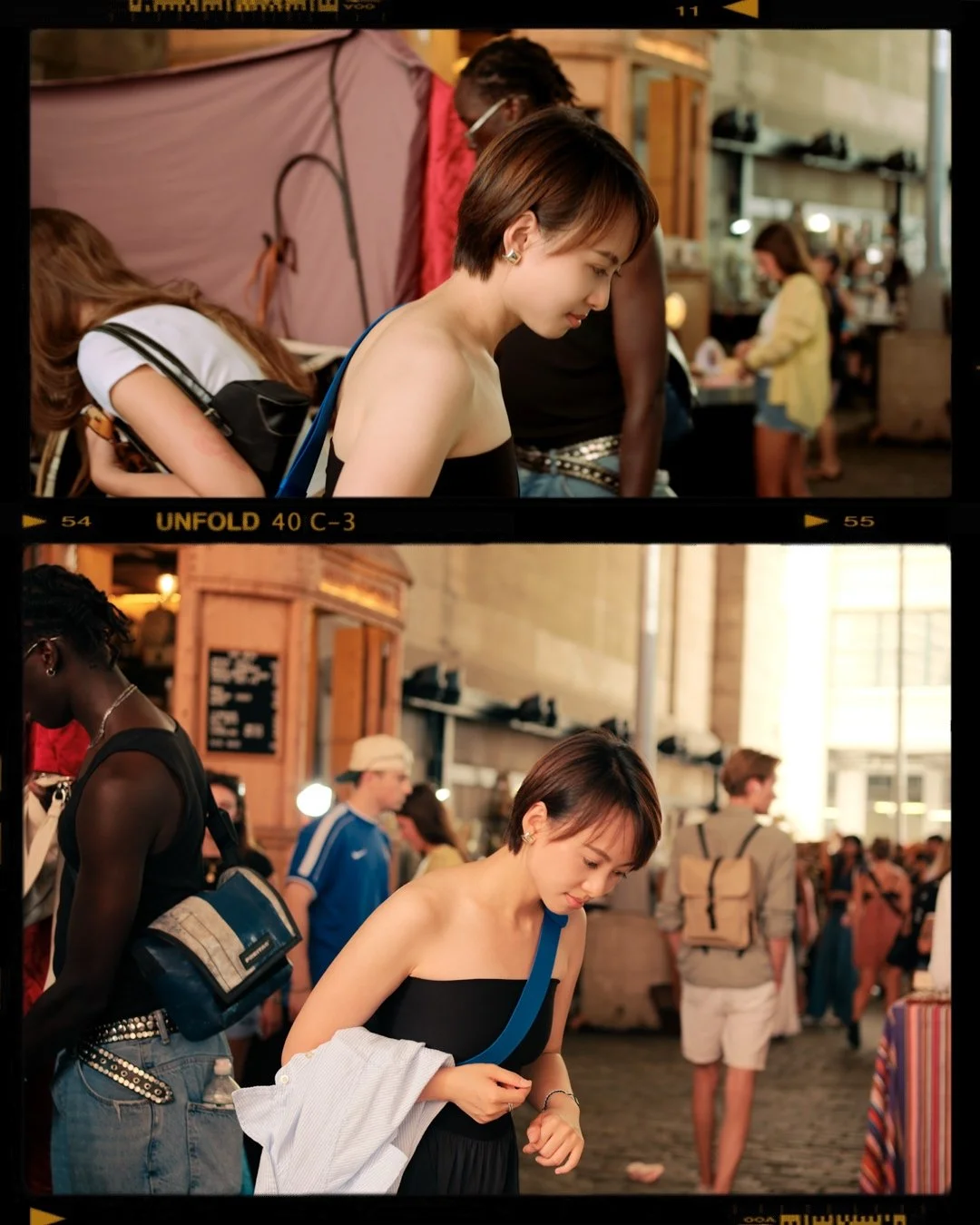 Young woman with short hair and a black strapless dress at a busy indoor market or train station, looking down thoughtfully, carrying a white jacket on her arm and a blue strap bag.