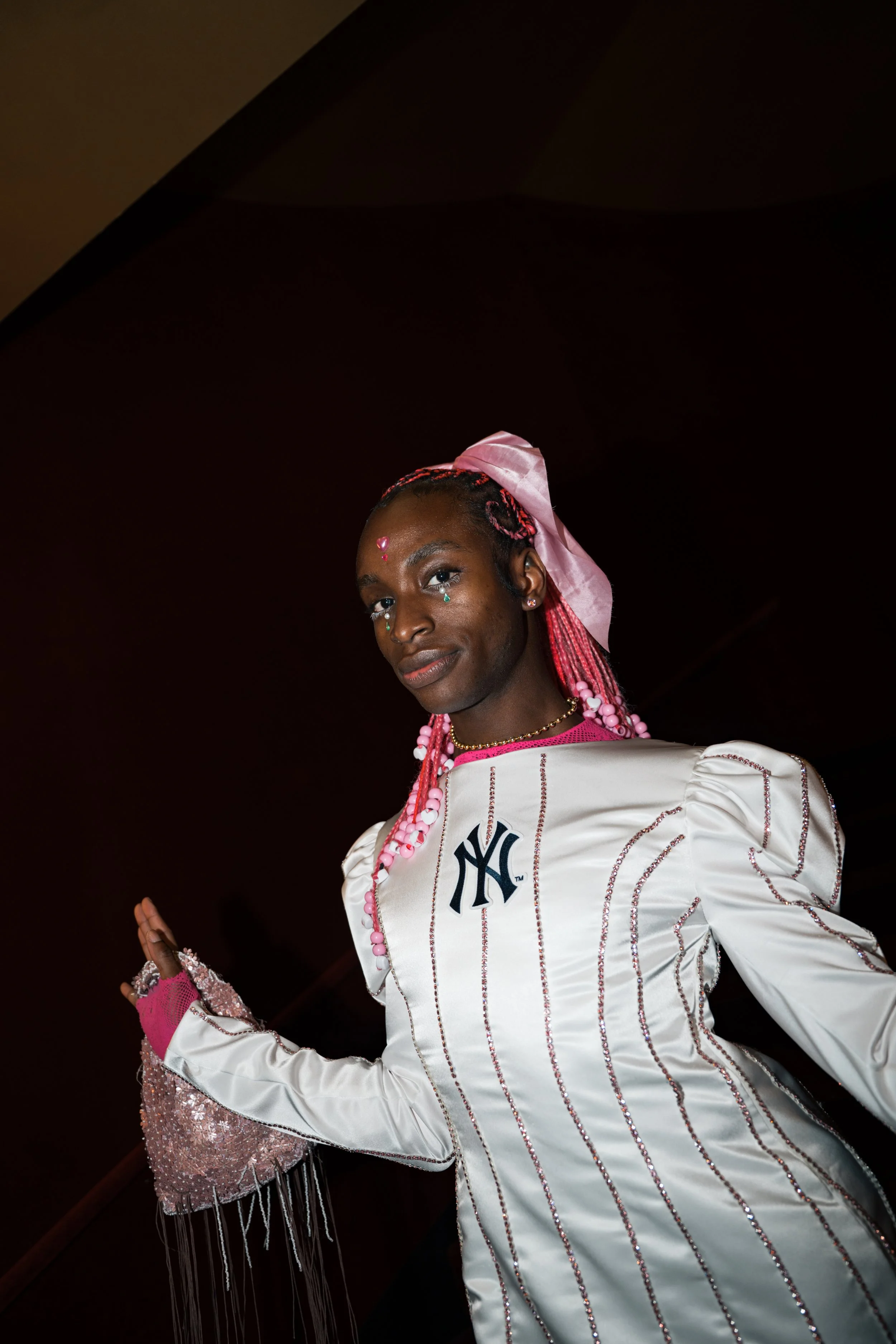 Person wearing a white NY Yankees jacket with pink accents, pink hair accessories, and decorative face stickers, standing against a dark background.