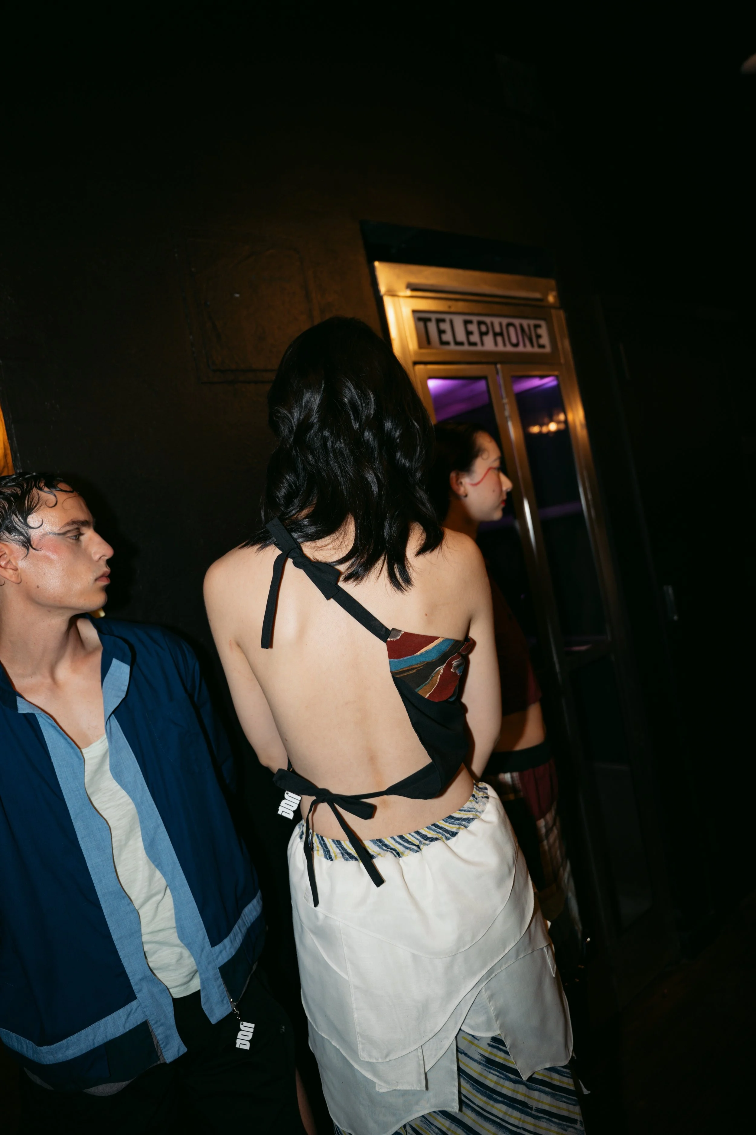 People standing in line near a telephone booth in a dimly lit indoor setting.
