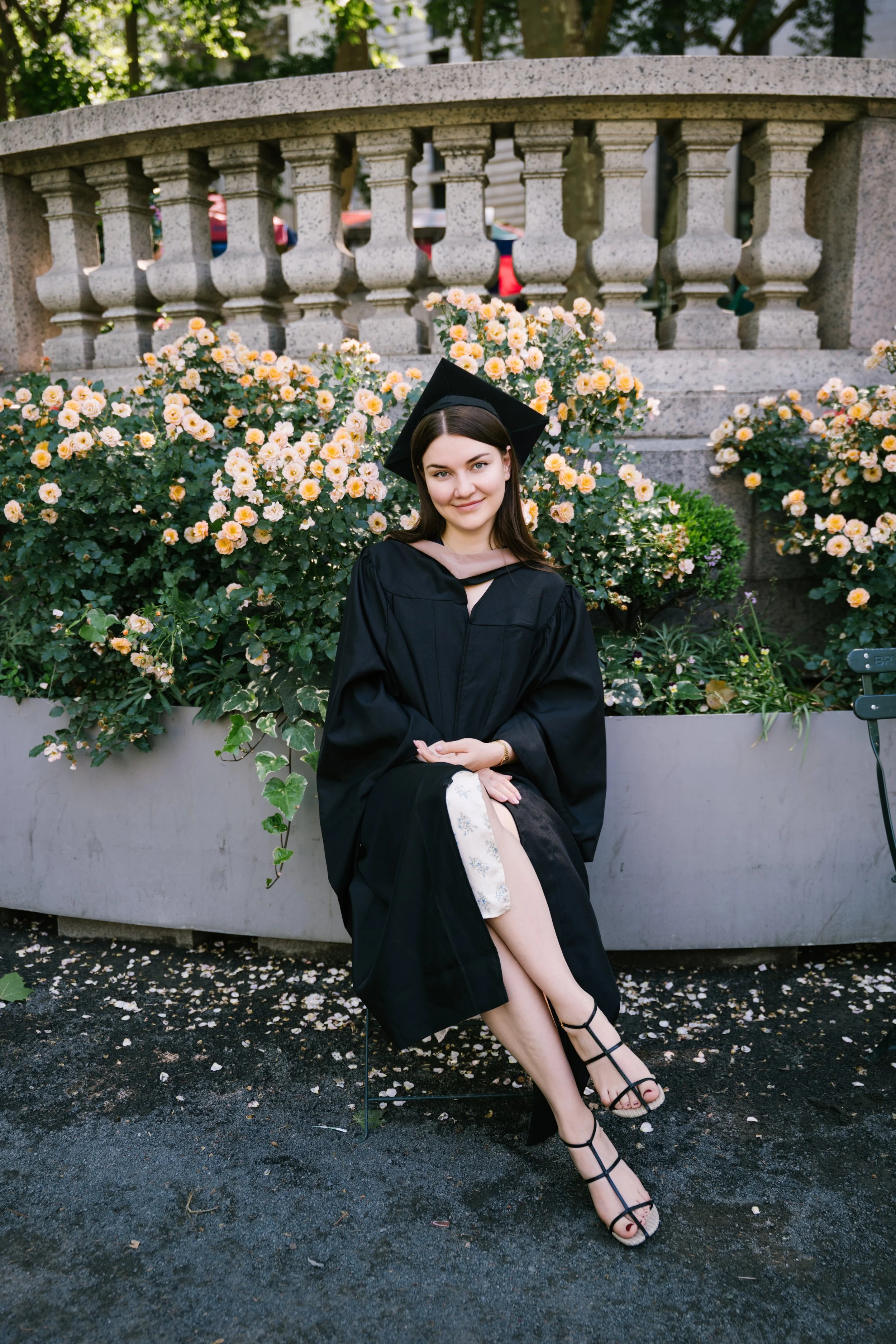 A young woman in graduation cap and gown sitting outdoors in front of a stone railing with yellow and pink flowers, smiling and crossing her legs, wearing black high-heeled sandals.