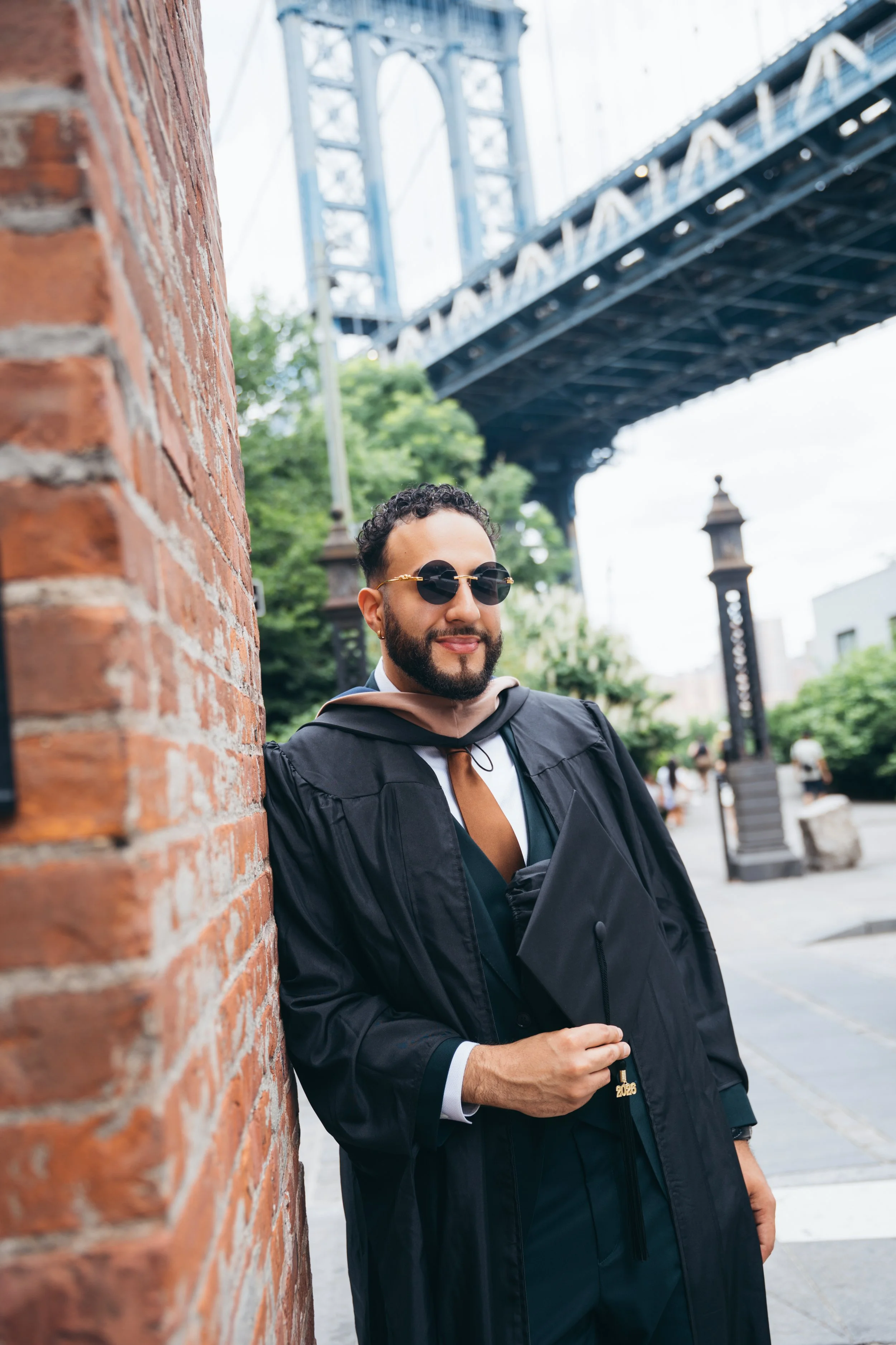 A man in graduation gown and cap leaning against a brick wall under a bridge with green trees in the background.