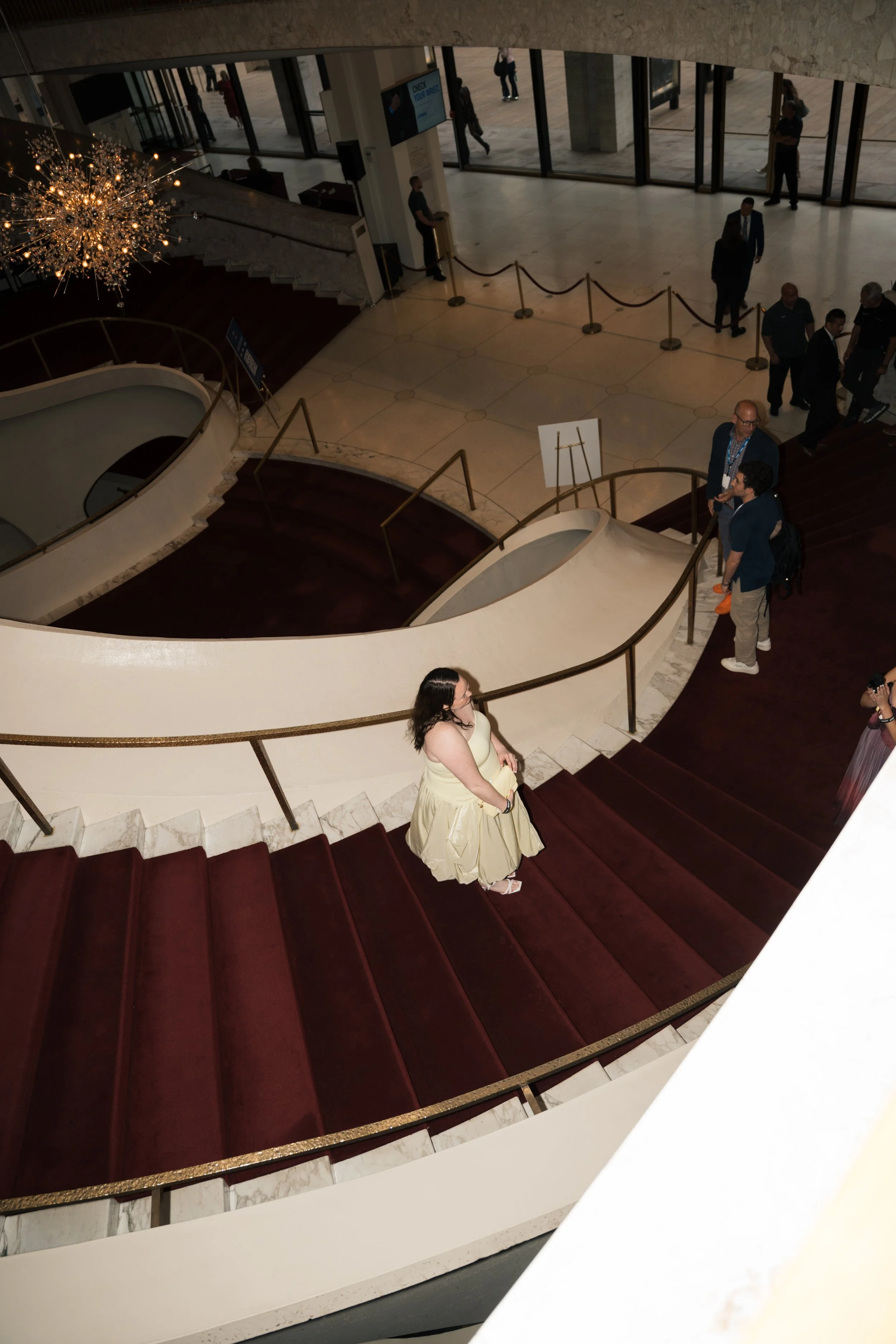 View from the top of a grand staircase inside a building, showing a woman in a yellow gown descending the stairs, with several people at the entrance and on the stairs, a chandelier hanging from the ceiling, and large glass doors letting in natural l