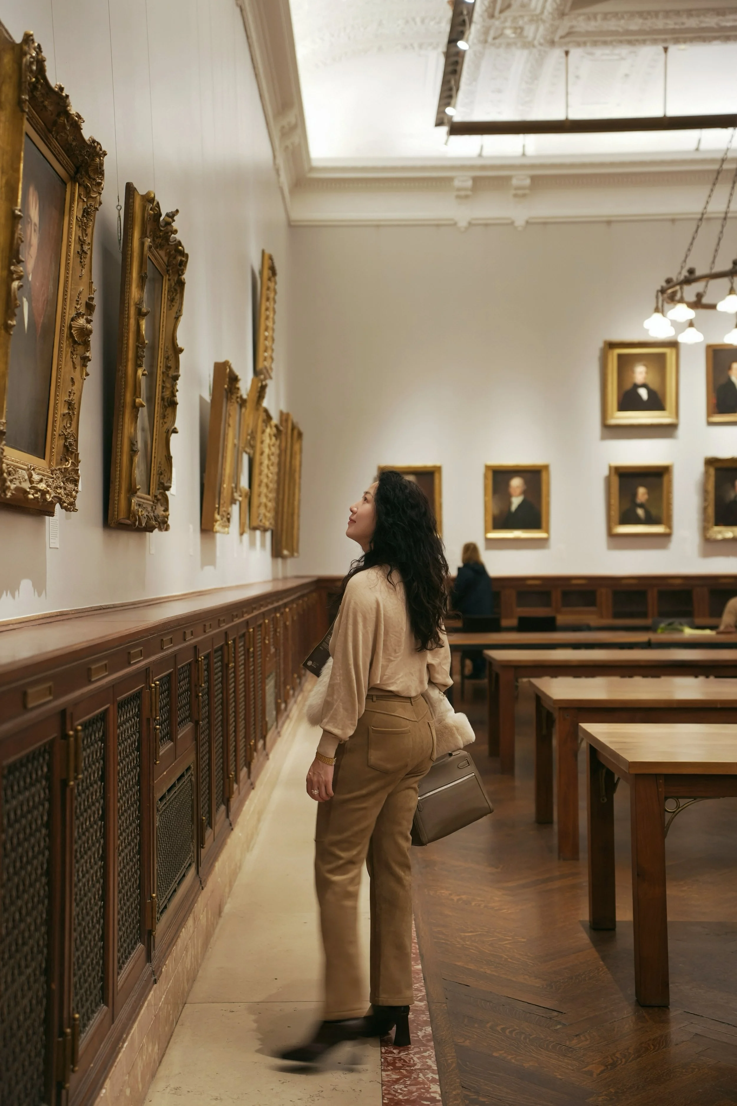A woman with dark, curly hair looking at framed portraits in an art gallery or museum, with wooden benches and a high ceiling.
