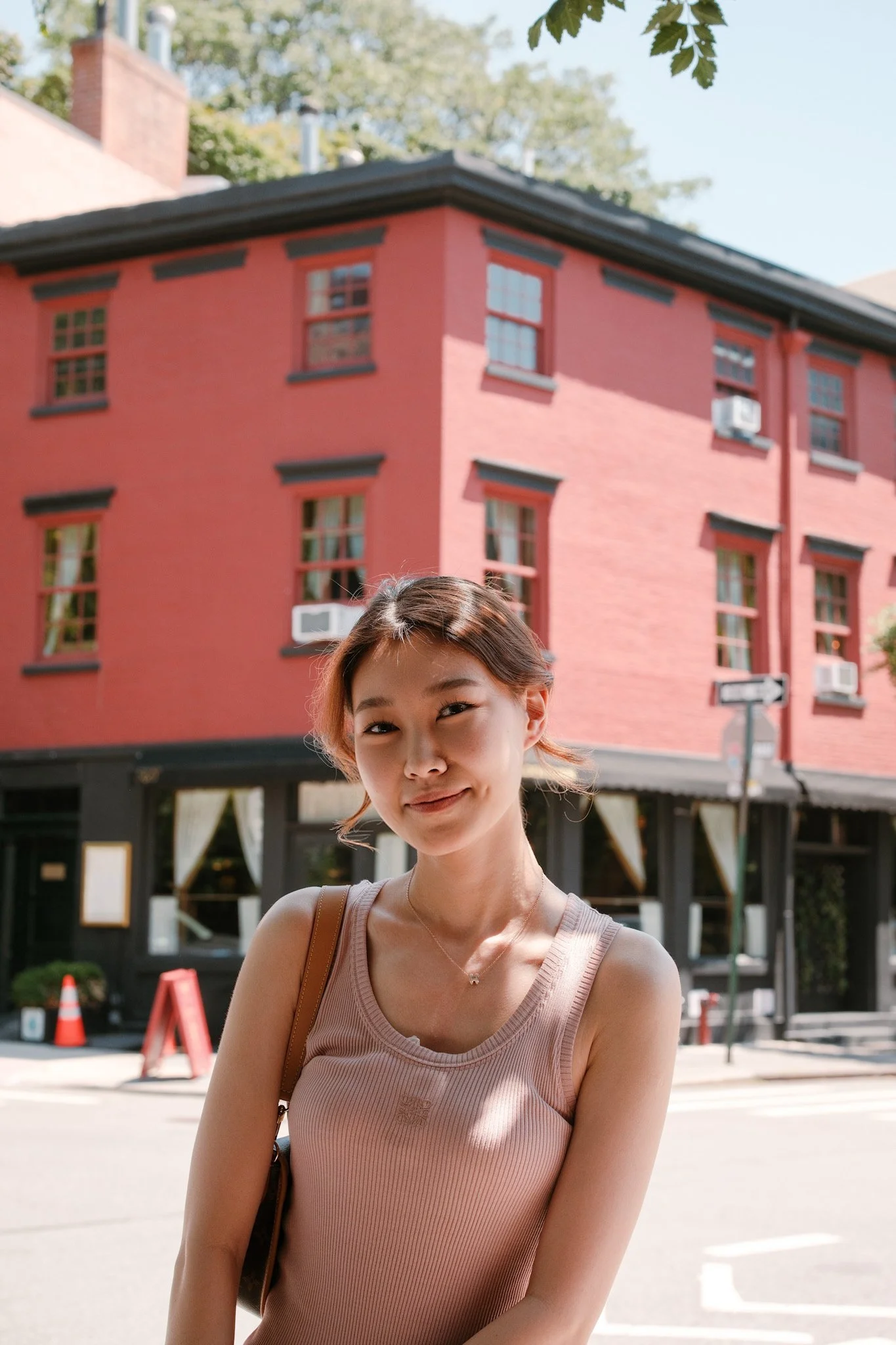 A smiling woman with short, light brown hair standing outdoors in front of a red brick building with multiple windows and air conditioning units. She is wearing a sleeveless beige top and a small necklace.