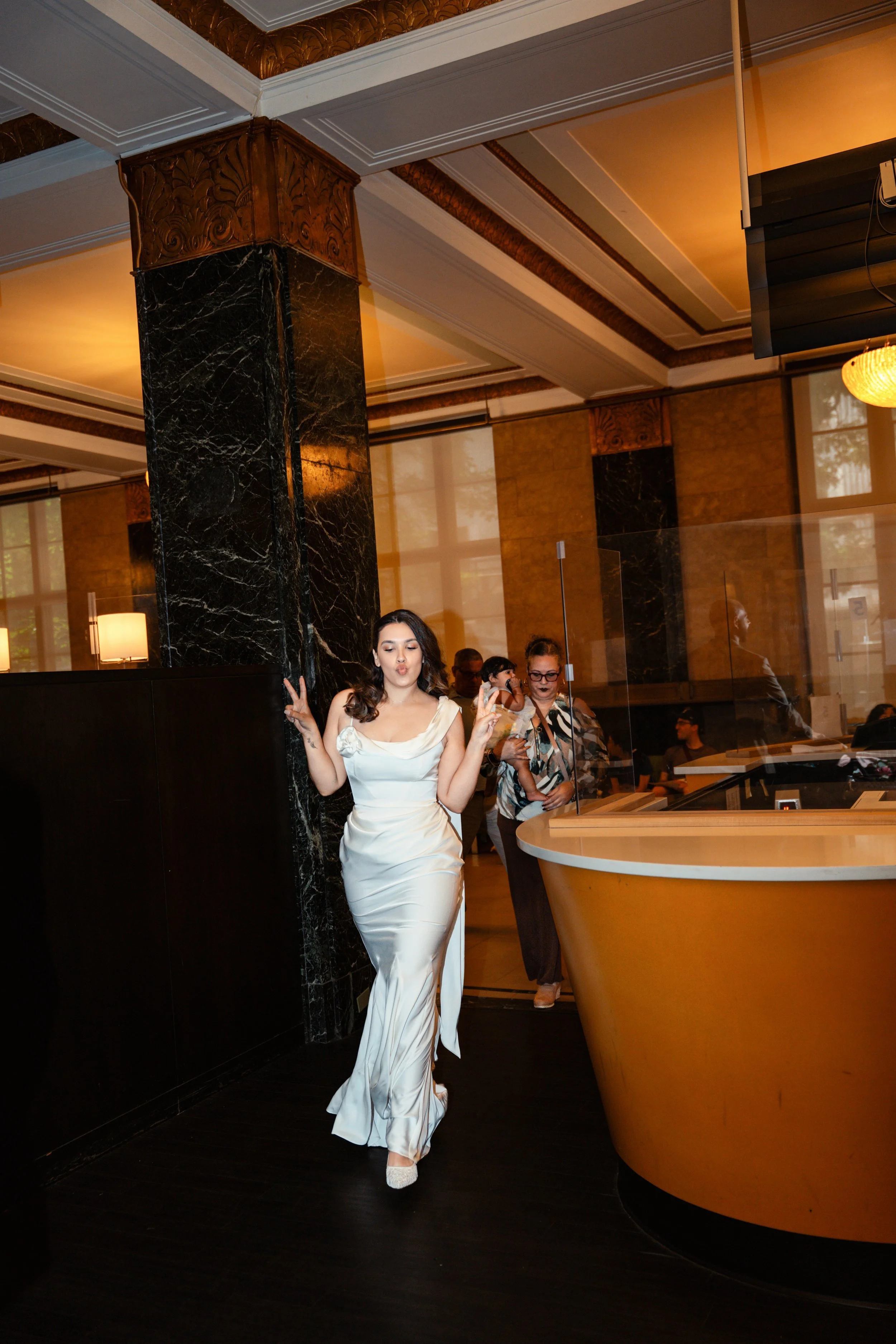 A woman in a white satin gown with a slit poses making peace signs at an indoor event in a hotel lobby with warm lighting, marble columns, and gold accents.