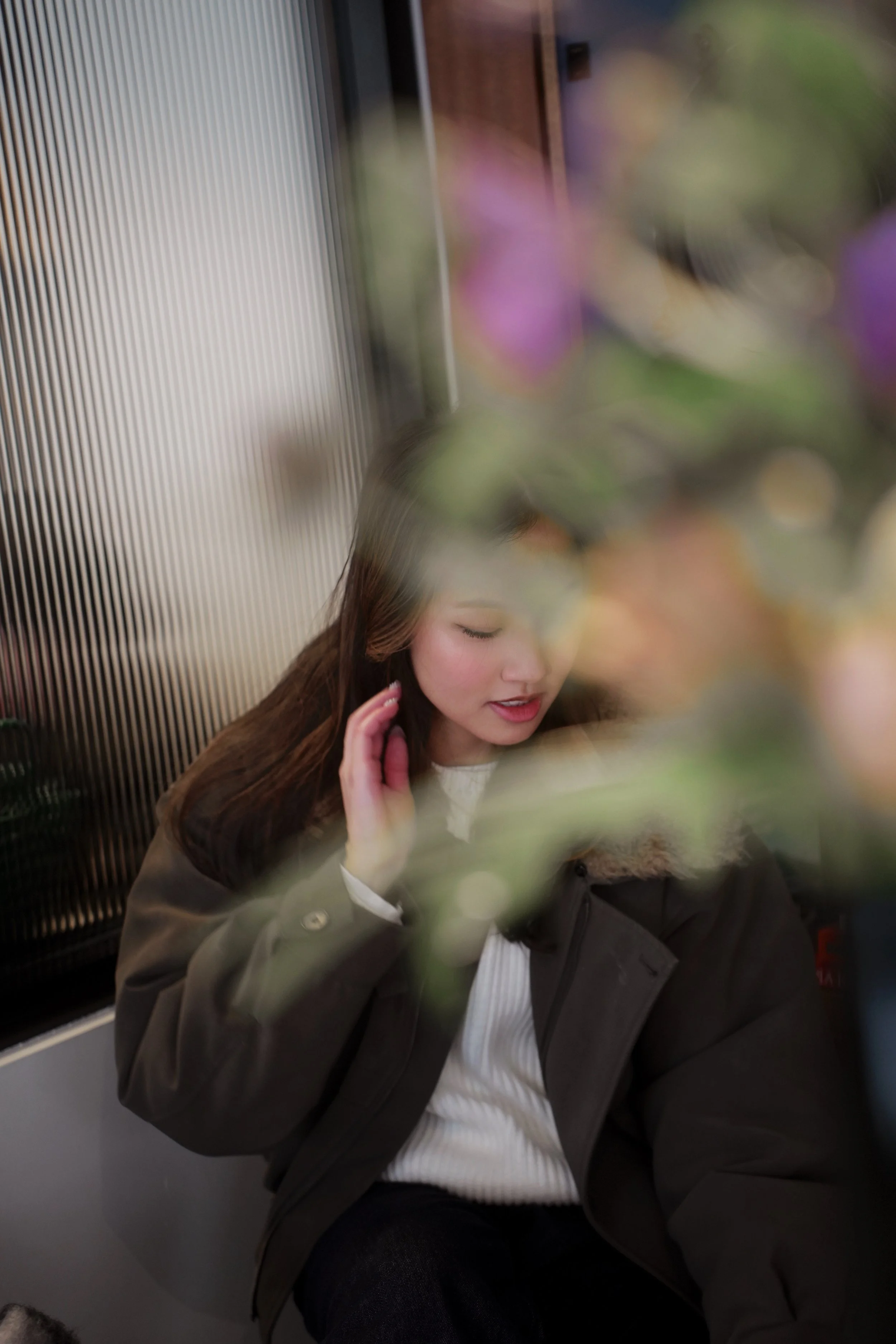A young woman with long brown hair and light skin, wearing a brown jacket and white top, sitting in an elevator with a metallic wall, her eyes closed and hand near face, partially obscured by a large blurry flower in the foreground.