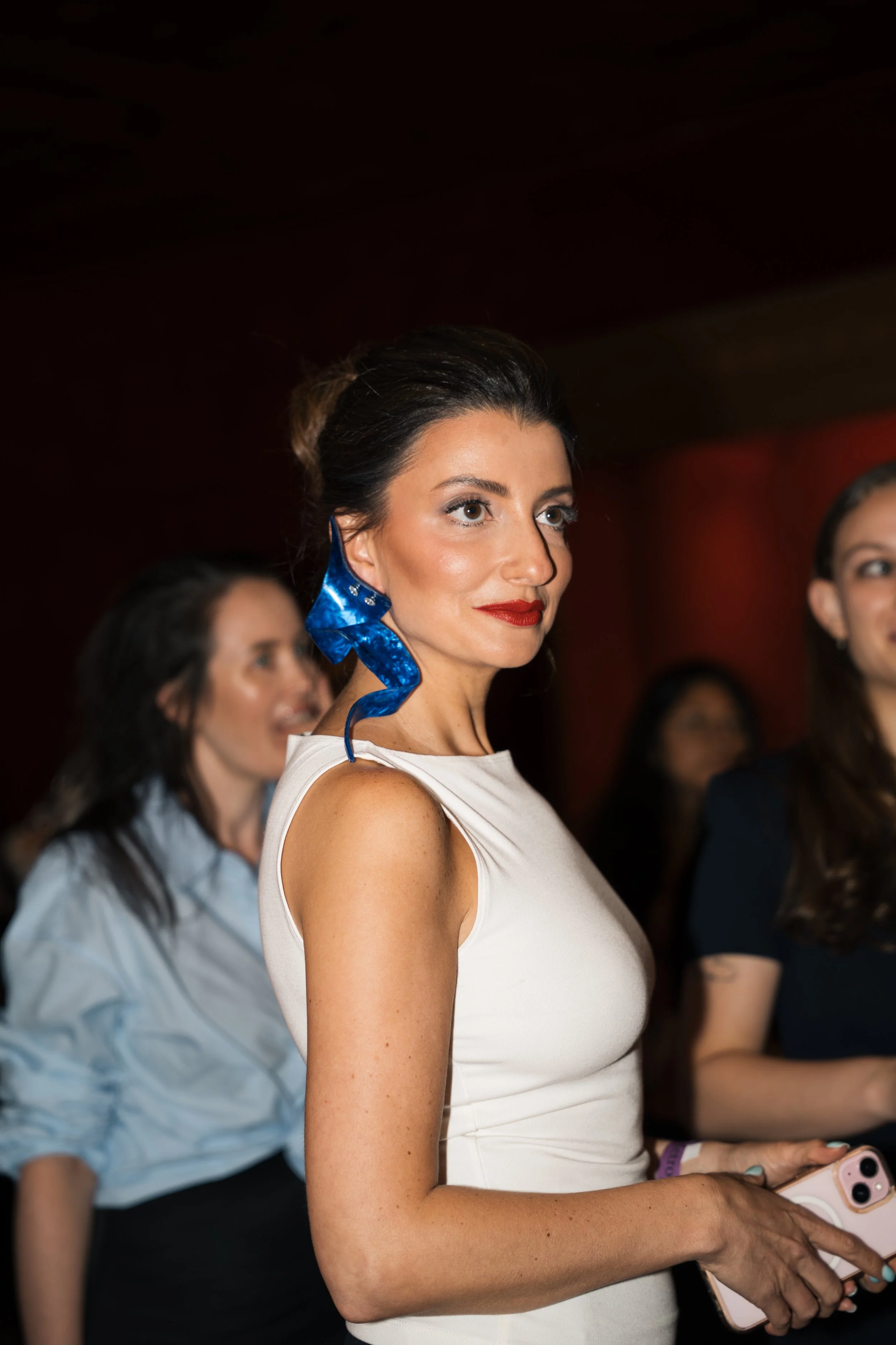 A woman with dark hair styled in an updo wearing a white sleeveless top holds a smartphone, with women sitting behind her in a dimly lit setting.