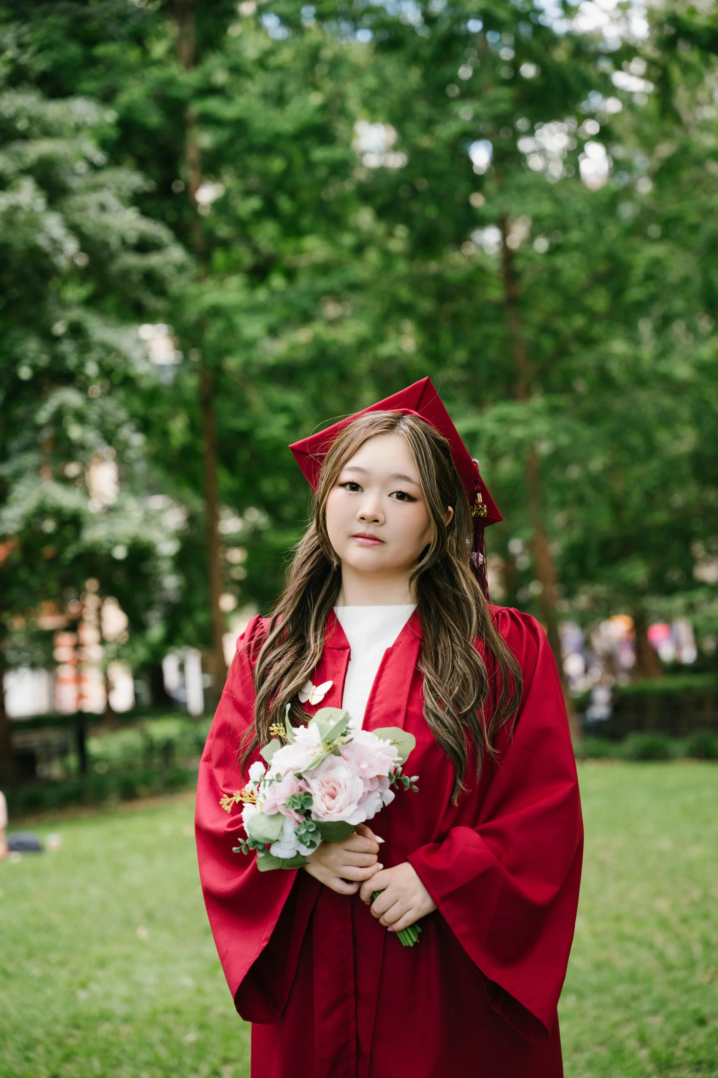 A young woman in a red graduation gown and cap with a gold tassel, holding a bouquet of pink and white flowers, standing outdoors in a park with green trees in the background.