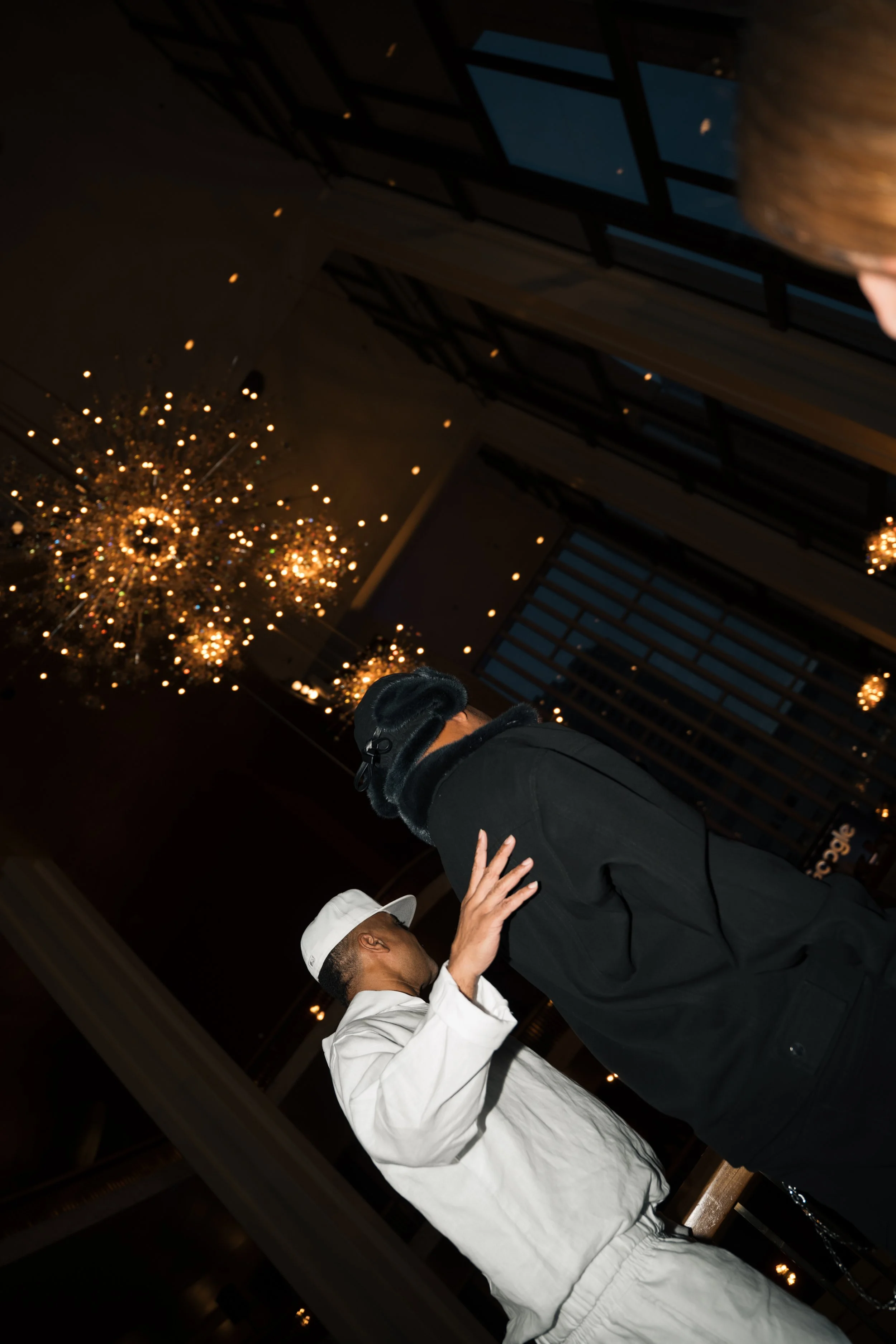 Two men, one in a white jacket and hat, the other in black with a chat gag, are dancing in a luxurious room with chandeliers and large windows.