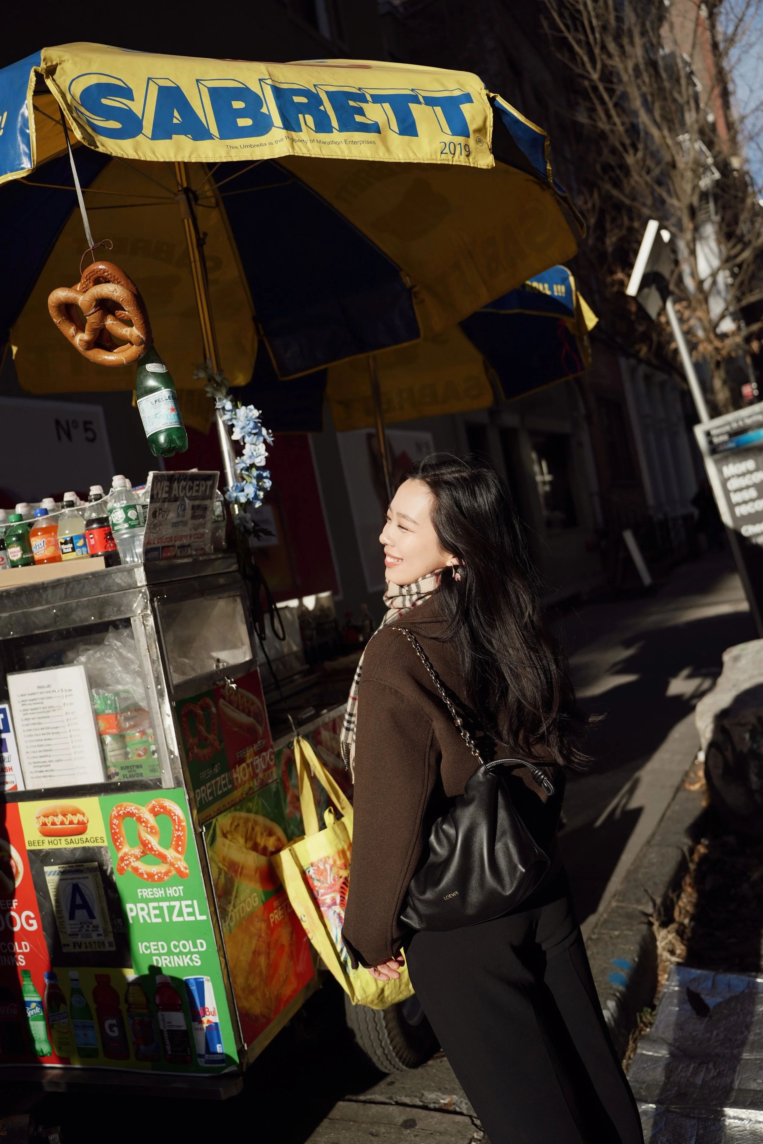 A young woman with long black hair, smiling with eyes closed, wearing a brown jacket and scarf, standing next to a hot dog stand on a city street in daylight. The stand has a yellow and blue umbrella and various snacks and drinks displayed.