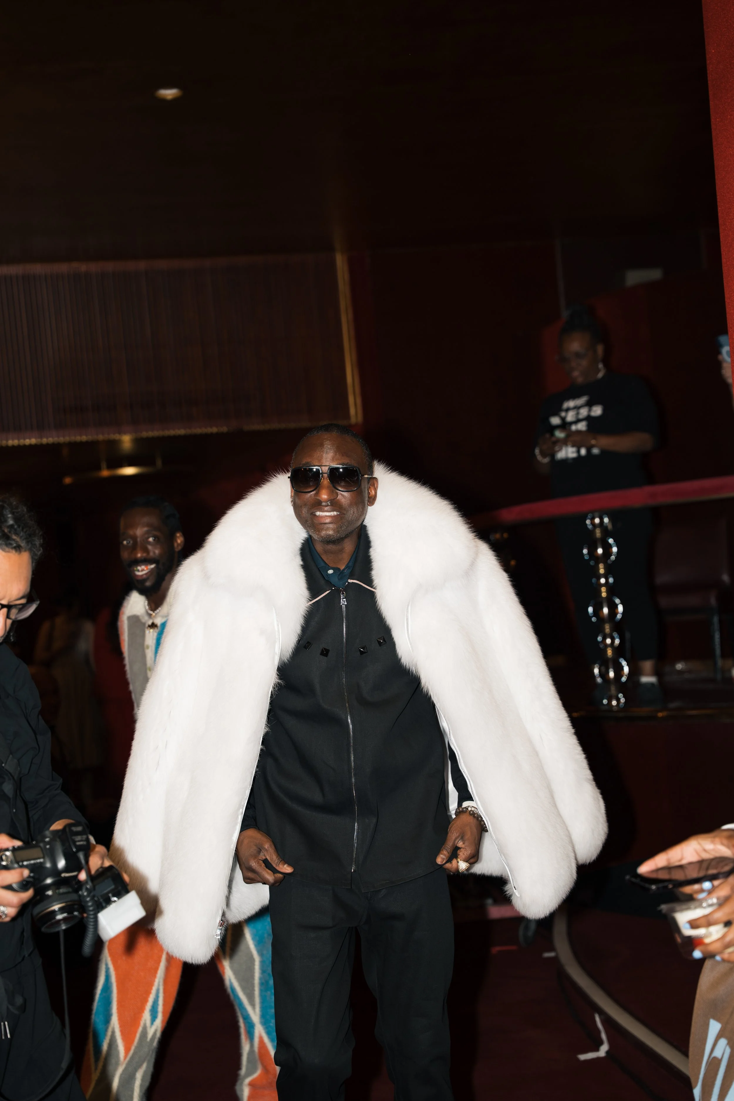 Man wearing a white fur coat, sunglasses, black shirt and black pants walking in an indoor setting with people around, some taking photos.