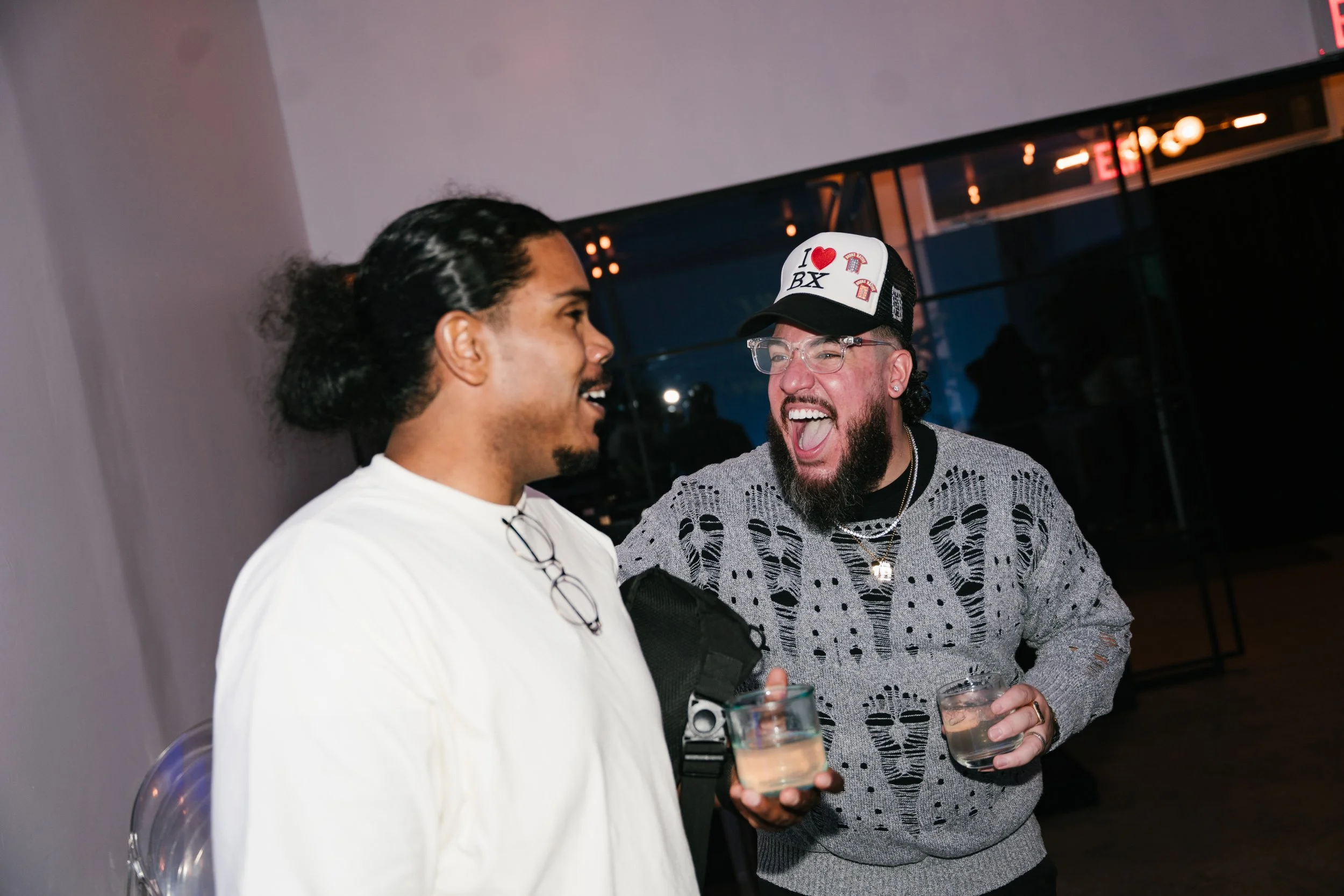 Two men laughing and holding glasses of beverages at an indoor event, one wearing a white shirt with glasses hanging on his neck, the other wearing a patterned shirt and a trucker hat with a heart and t-shirt image, in front of a dark background with
