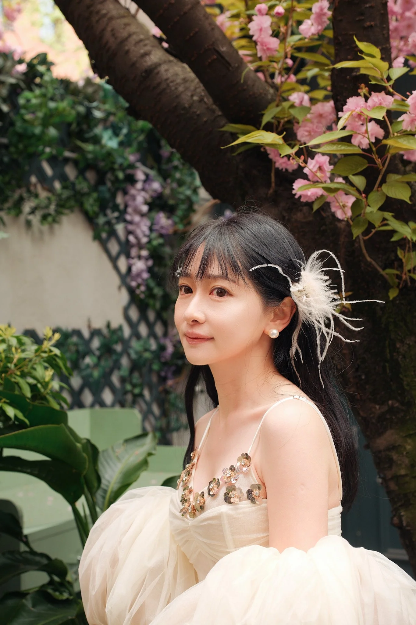 A woman with long black hair and pearl earrings is wearing a cream-colored dress with a decorated bodice and a large, puffy skirt. She is standing outdoors in front of a background of pink cherry blossoms and green foliage.