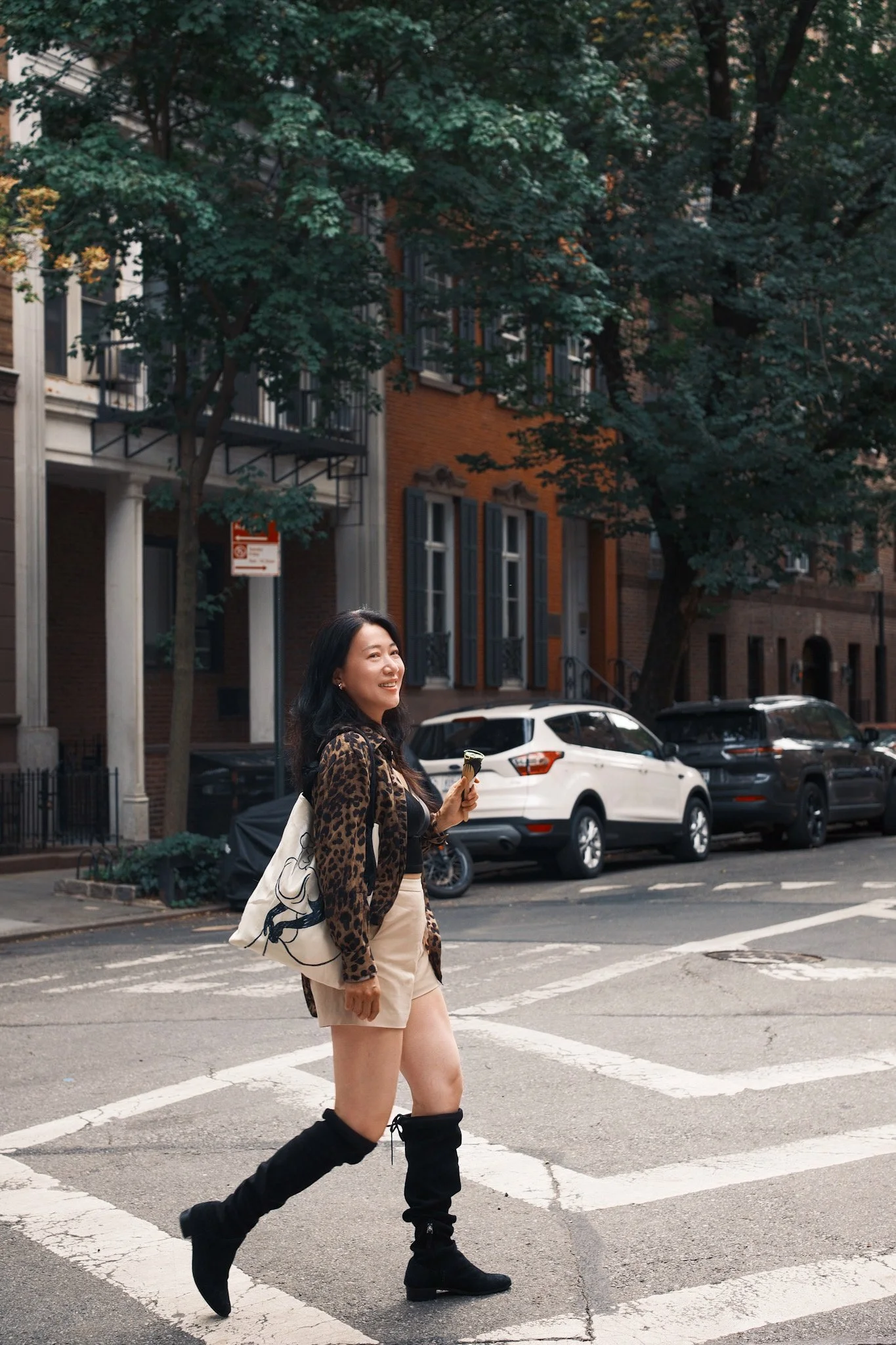 A woman with long dark hair wearing a leopard print jacket and beige shorts, crossing a street at a crosswalk, carrying a canvas bag, with a row of parked cars and brick buildings with green trees in the background.