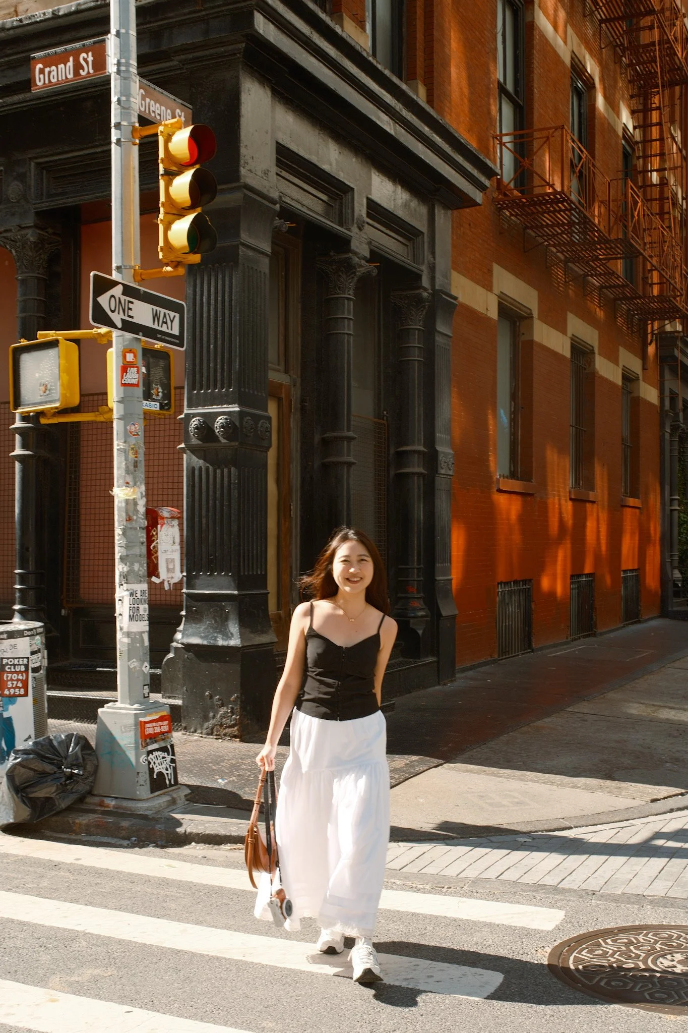 A young woman with long dark hair, wearing a black sleeveless top and white wide-leg pants, smiling and walking across a city crosswalk. She is carrying a brown handbag and a small object in her right hand. The background features a red brick buildin