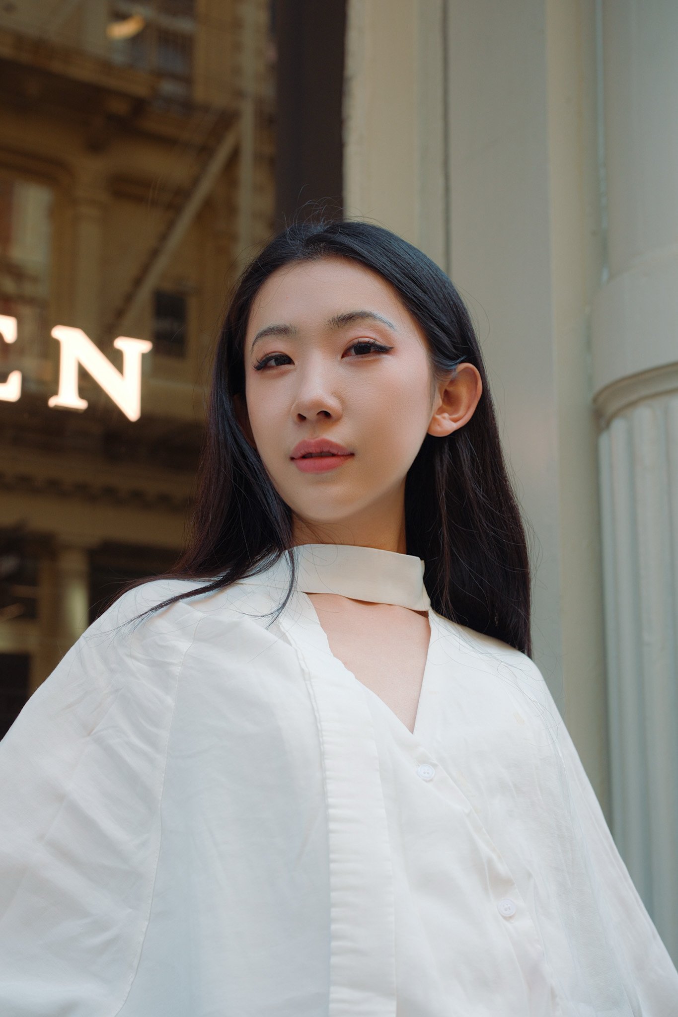 A young woman with long black hair wearing a white blouse, standing in a city street with a building and a sign in the background.
