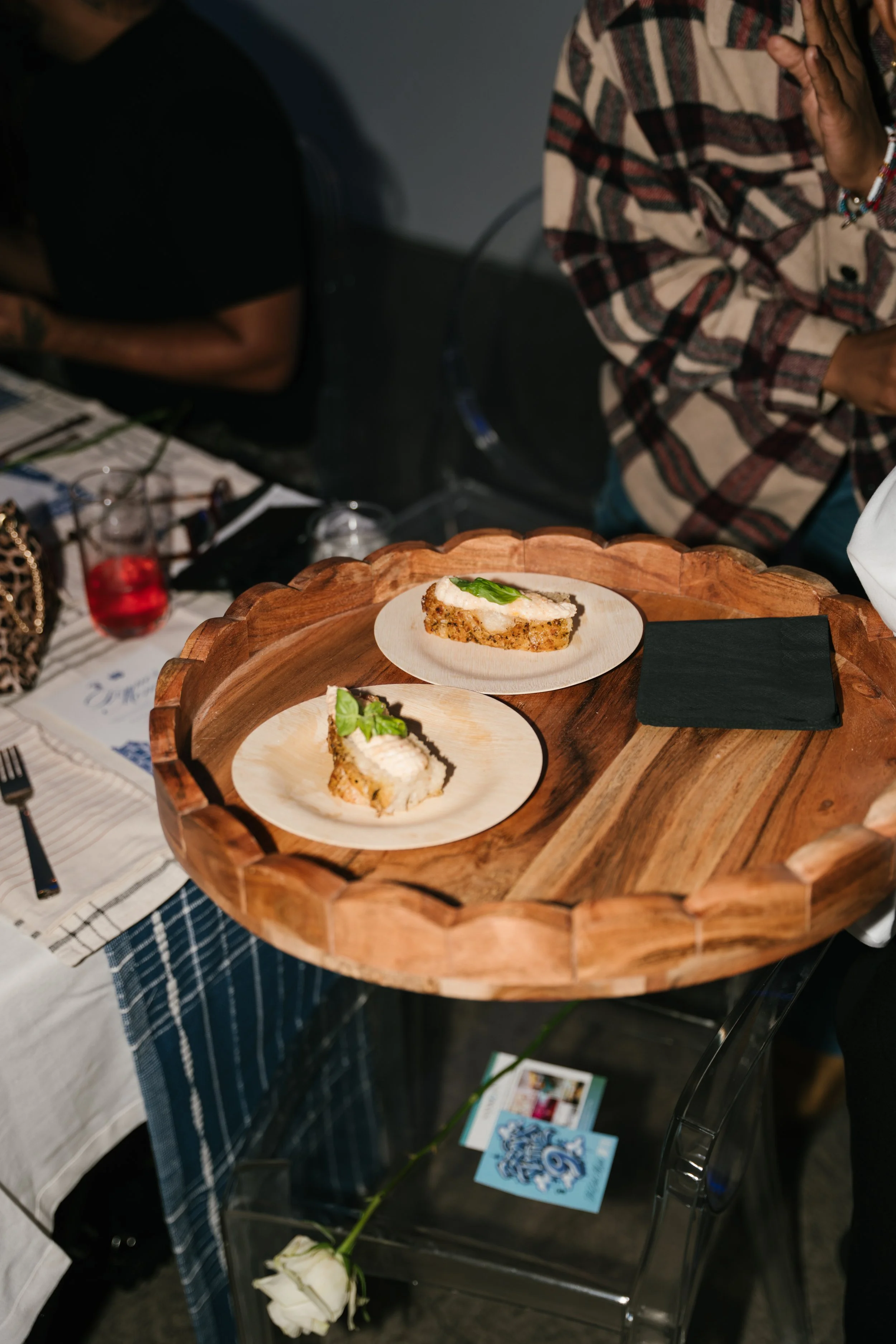 Plate with two pieces of cake, garnished with a green leaf, on a circular wooden tray at a table with a checkered tablecloth. A white rose and a brochure are also visible on the table.