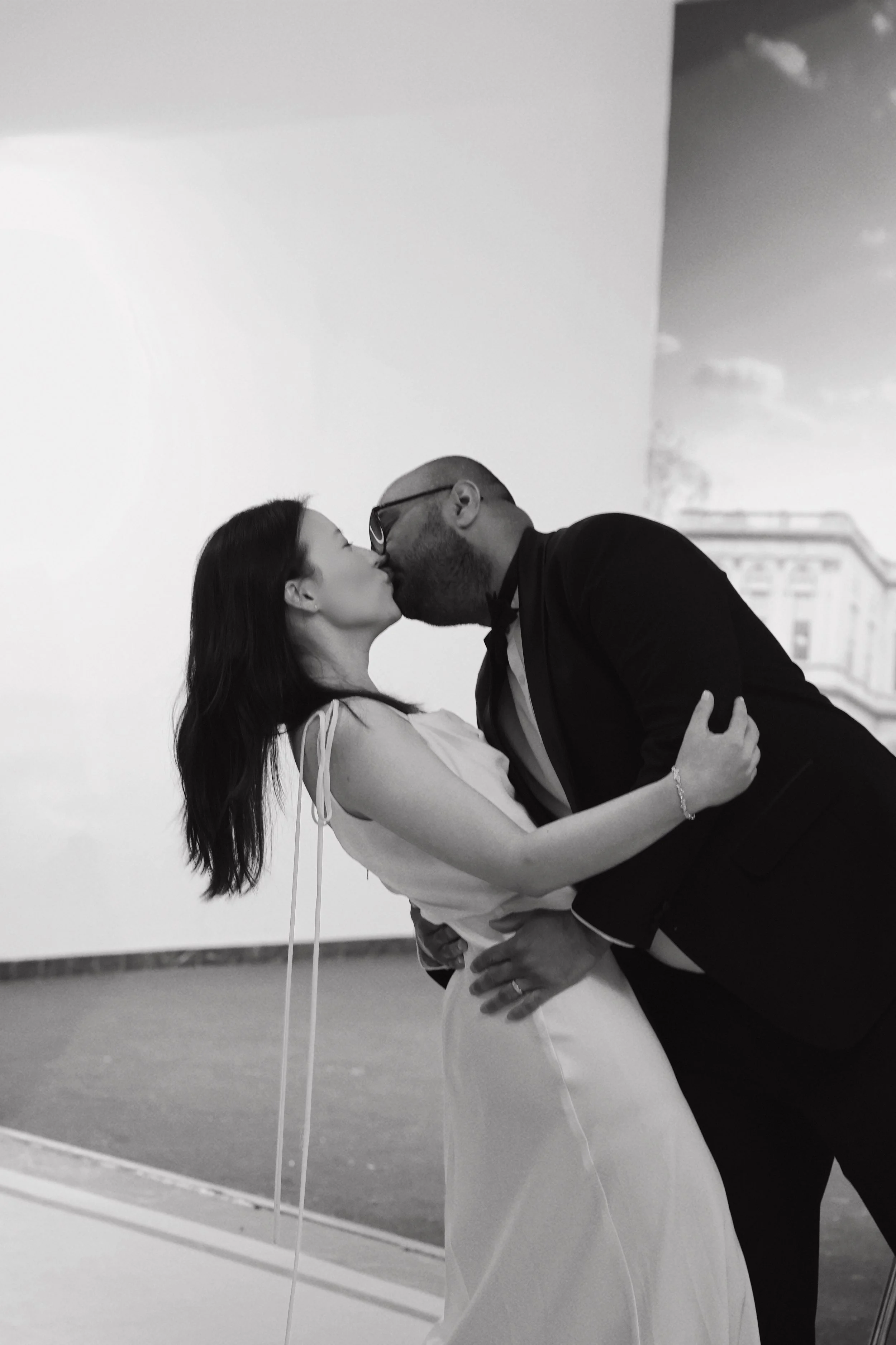 A couple dressed in formal attire sharing a dance, the man is dipping the woman, both smiling, in a room with a plain wall and a large mirror reflecting part of the scene.