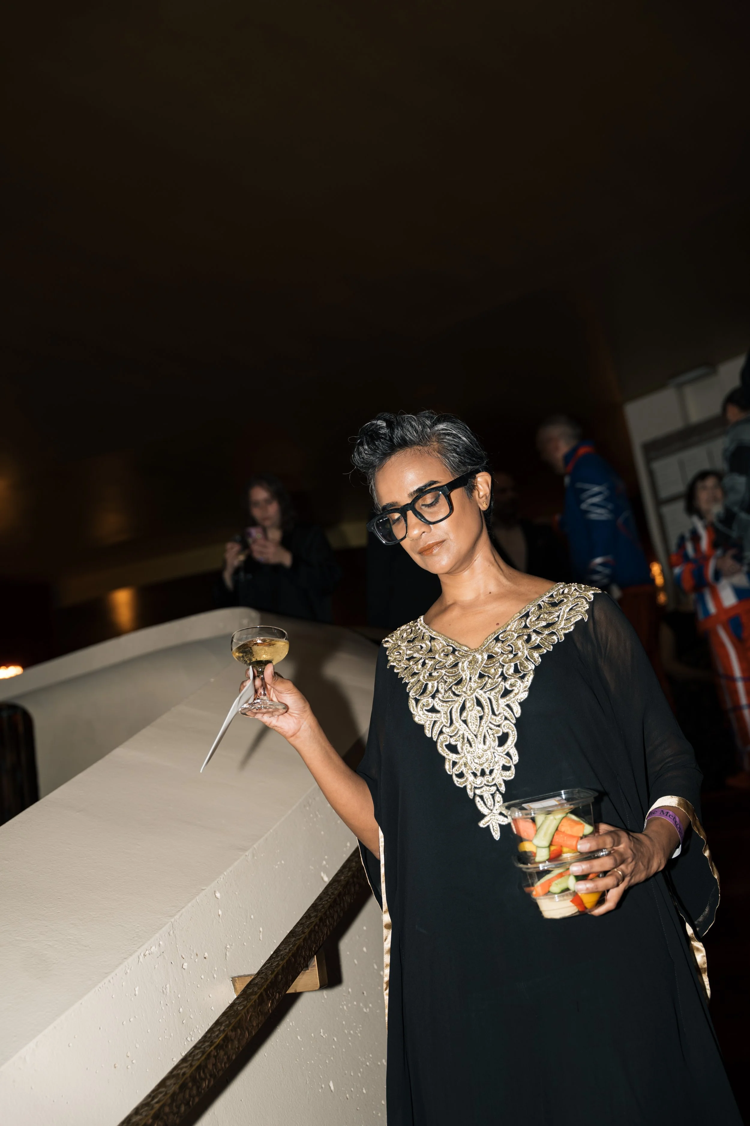 A woman with short dark hair, wearing glasses and a black dress with intricate gold embroidery, holds a glass of white wine in one hand and a bowl of mixed vegetables in the other, at a social event with other people in the background.