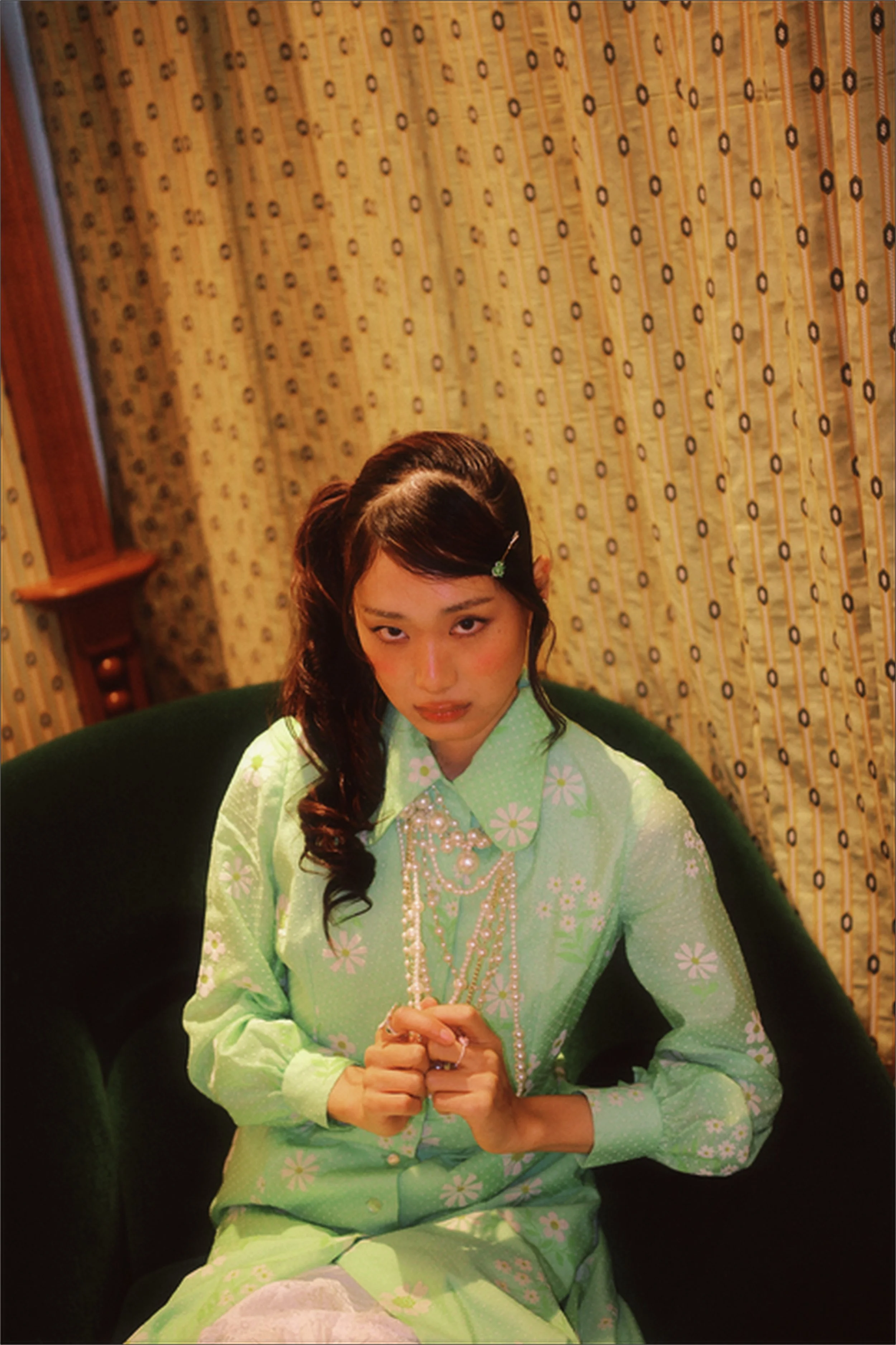 A woman in a bright green dress with a floral pattern, sitting on a dark green chair, with her hands clasped. She has long, dark, wavy hair with a small green clip, and is wearing layered necklaces. She is in a room with patterned yellow curtains and wooden furniture.