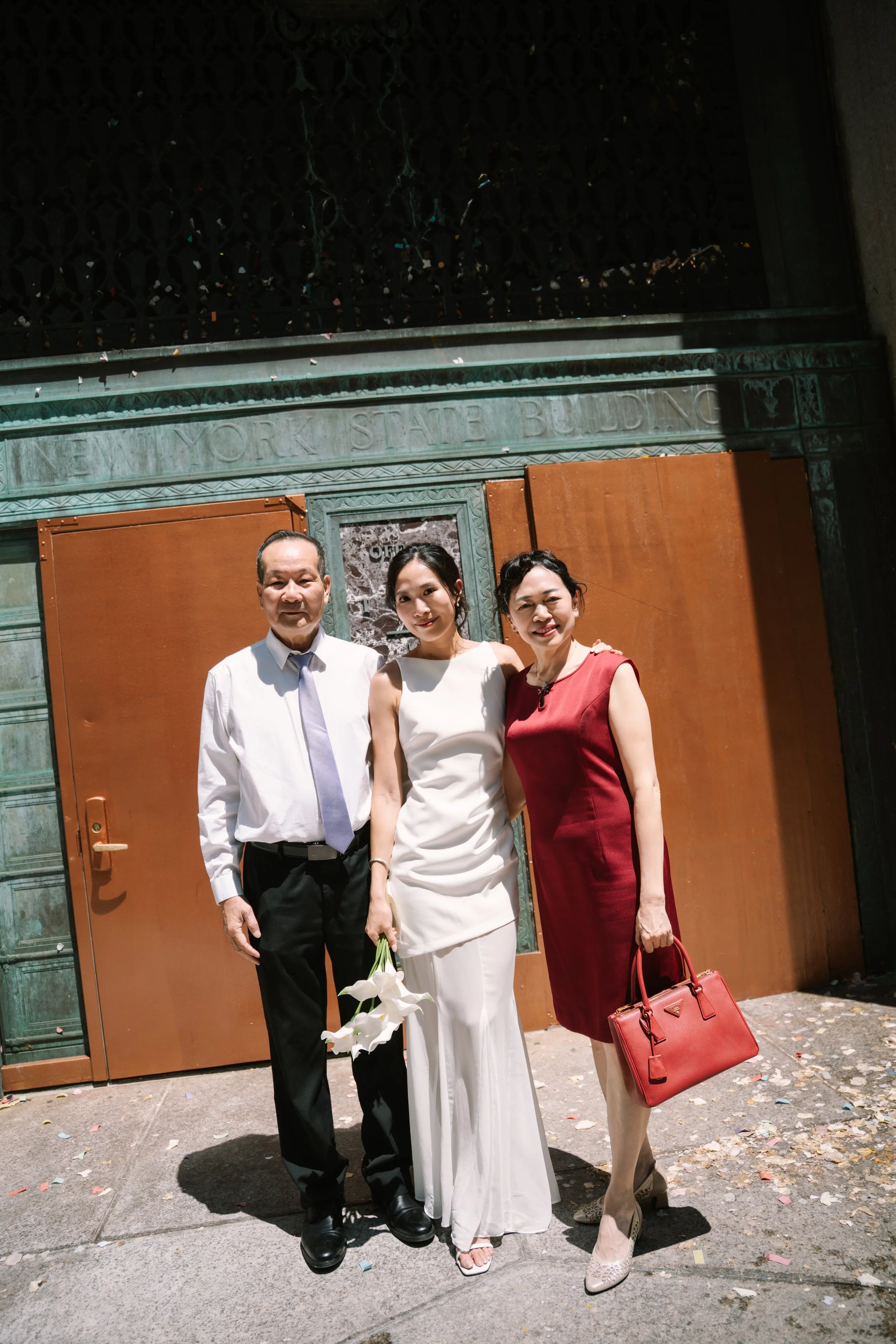 Three people standing outside in front of a building with a sign that reads 'New York State Building.' The person on the left is a man in a white shirt with a tie and black pants. The person in the middle is a woman in a white sleeveless dress holdin