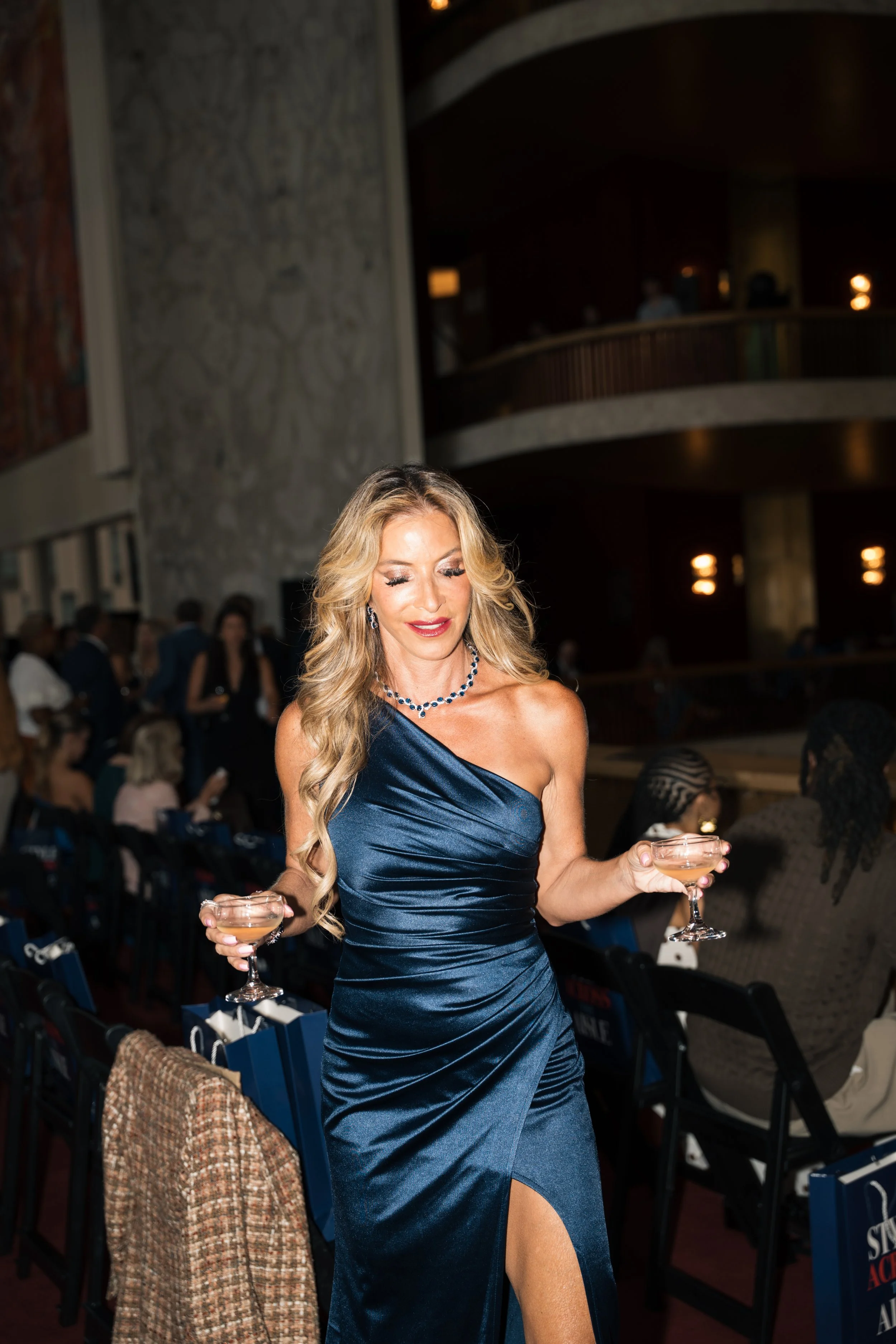 A woman in a blue satin evening gown with a thigh-high slit, holding a cocktail glass in each hand at an indoor event.