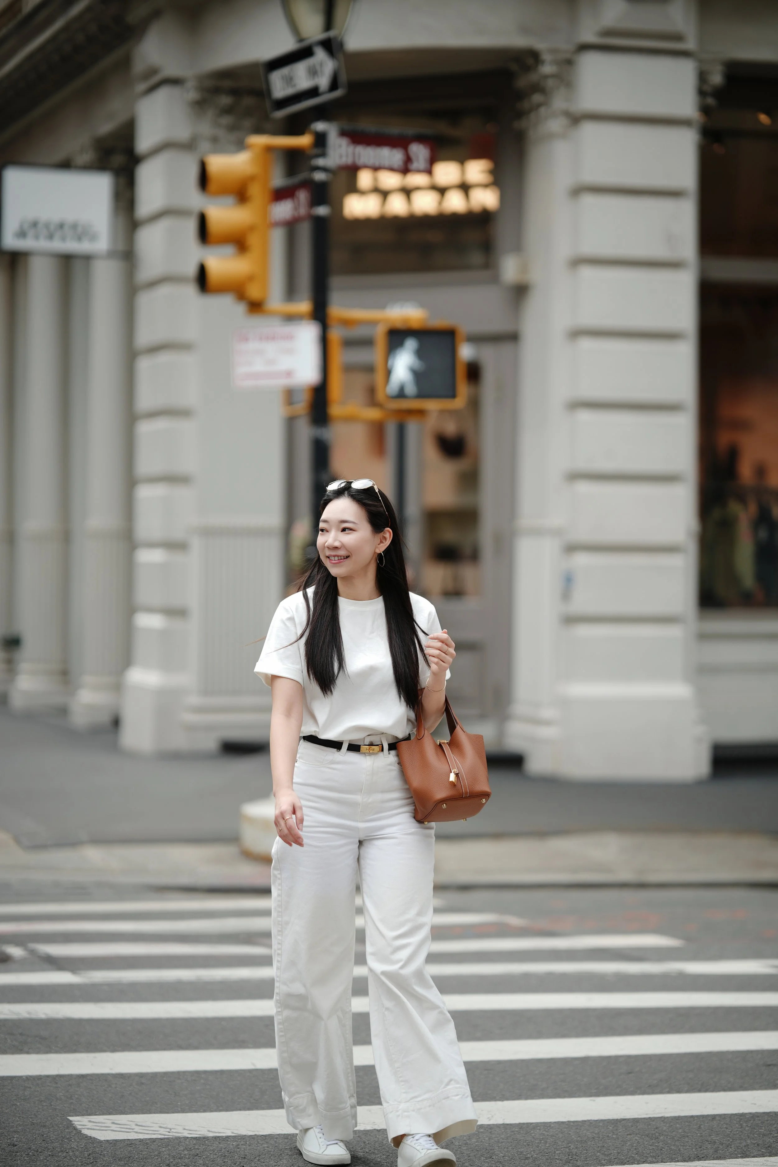 A young woman with long dark hair, dressed in white pants and a white t-shirt, stands on a crosswalk in an urban setting. She is smiling and holding a brown handbag over her shoulder, with sunglasses on her head. There are traffic lights and street s