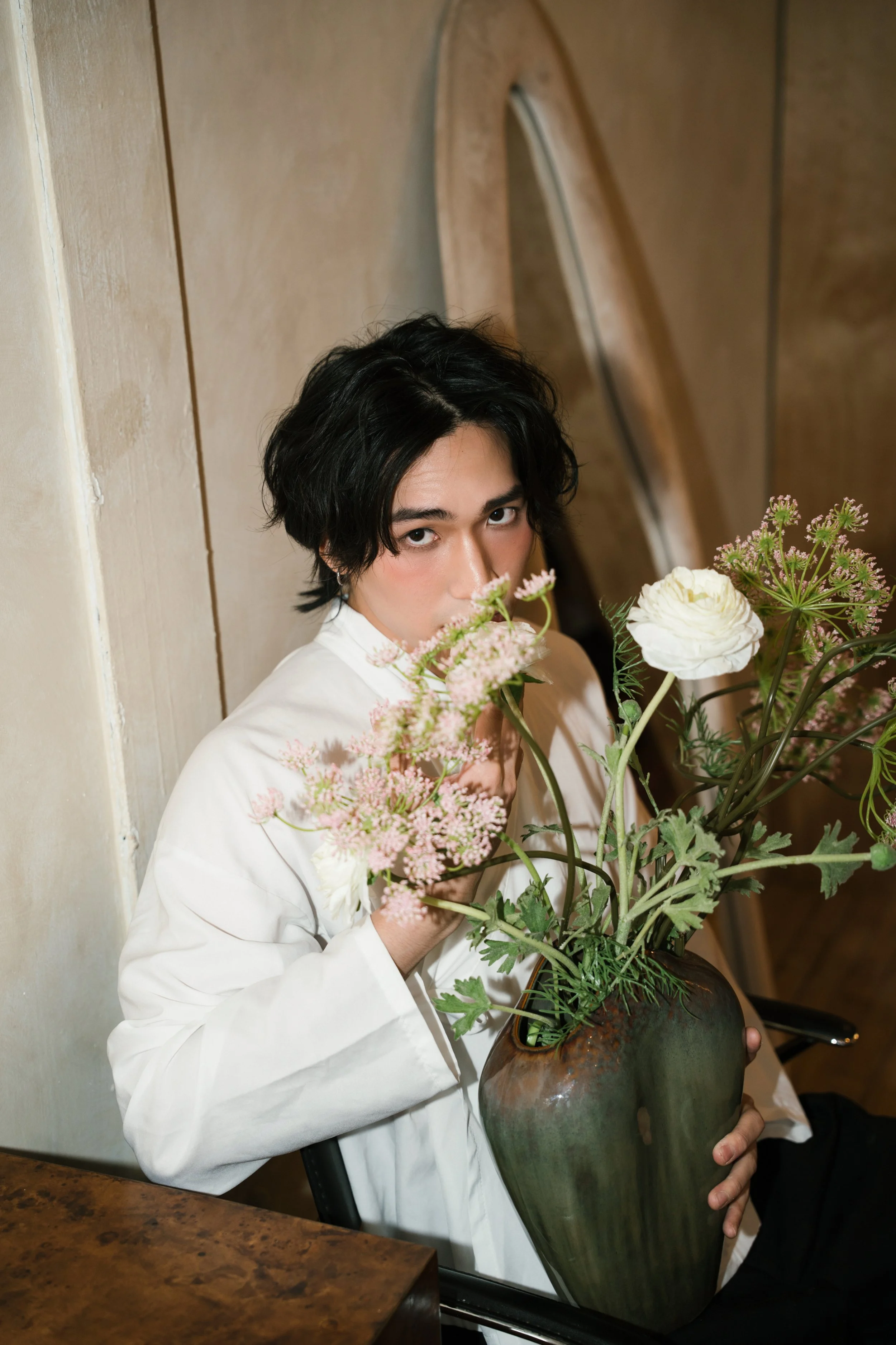 A person with black, wavy hair wearing a white shirt holding a large ceramic vase filled with pink and white flowers, looking directly at the camera in an indoor setting.