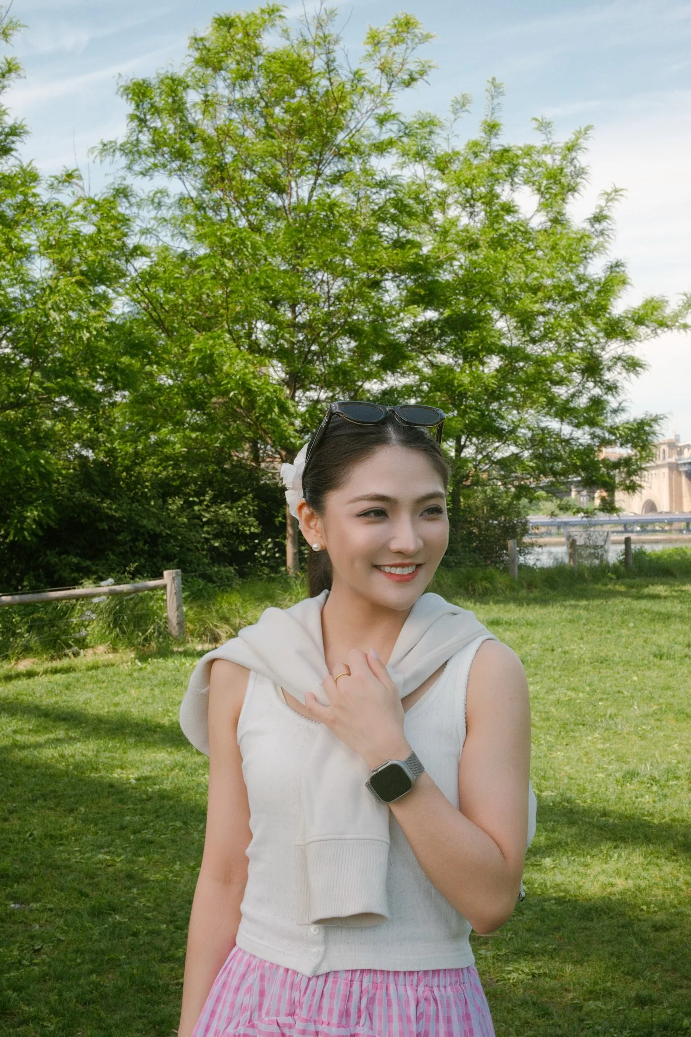 A young woman with dark hair and sunglasses on her head smiles outdoors in a park with trees and a building in the background. She is wearing a white sleeveless top, a pink and white striped skirt, and a smartwatch on her left wrist. She is holding a
