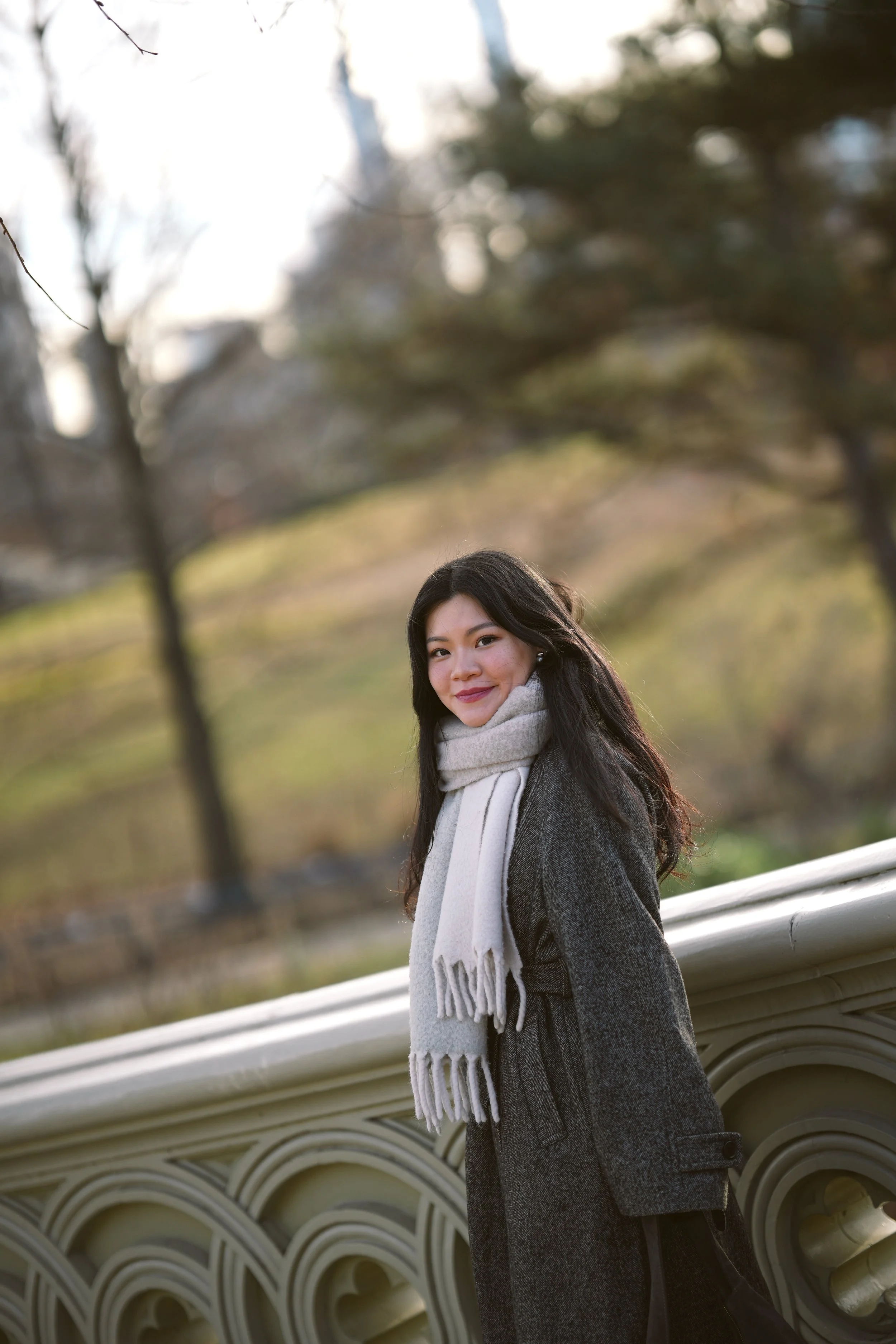 A woman with long dark hair smiling outdoors in a park, wearing a dark coat and a light-colored scarf, standing next to a decorative white railing with trees in the background.