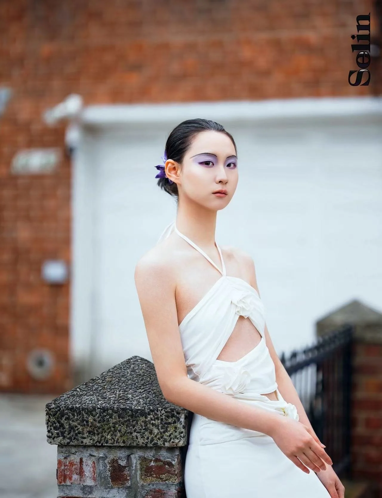 A woman with dark hair styled back, wearing pale purple eyeshadow, a white halter dress with ruffled details, sitting outdoors against a brick wall and white background, leaning on a stone and brick pillar.