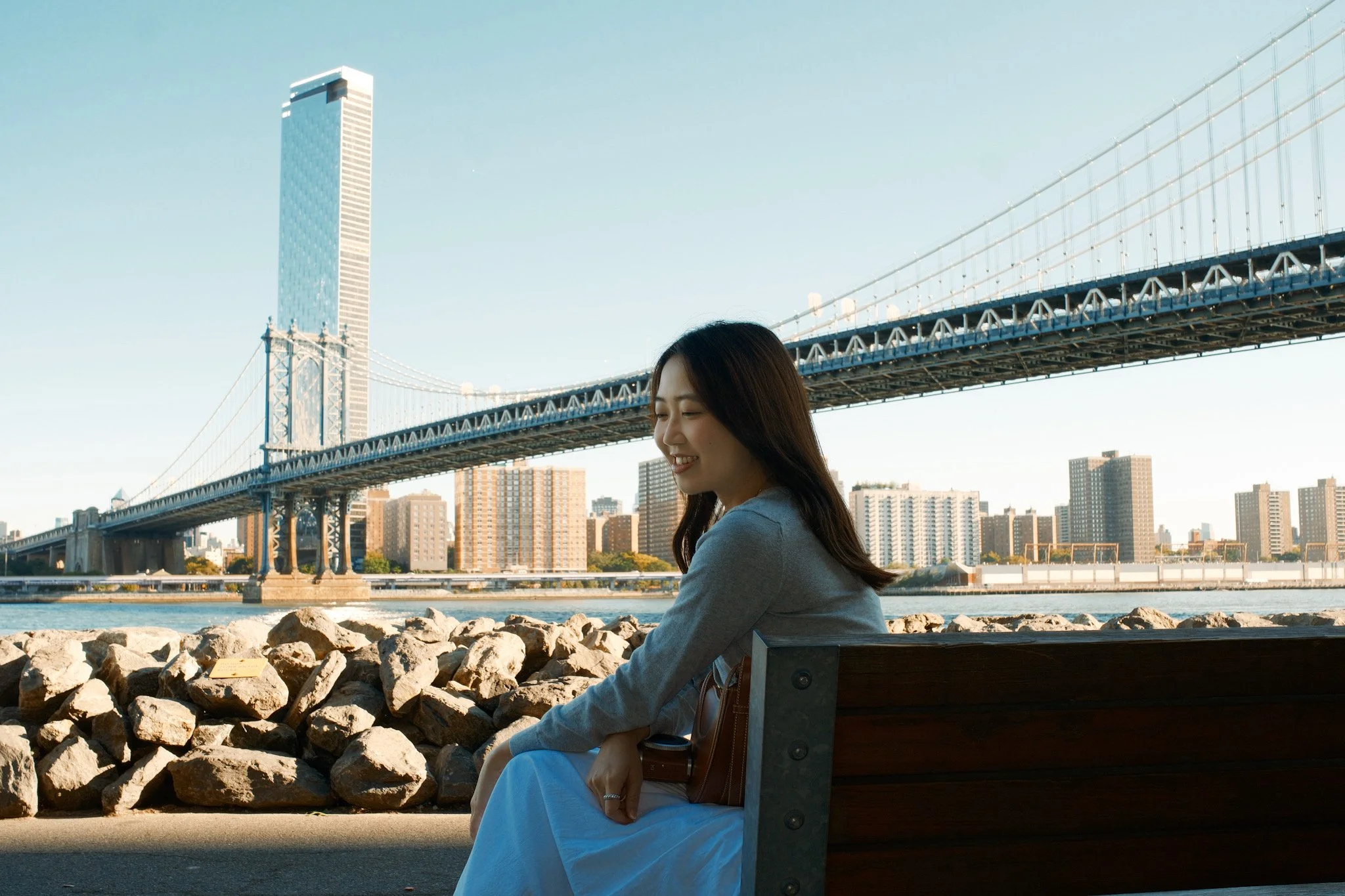 A woman sitting on a park bench near a river, with a city skyline and a suspension bridge in the background, smiling with her eyes closed.