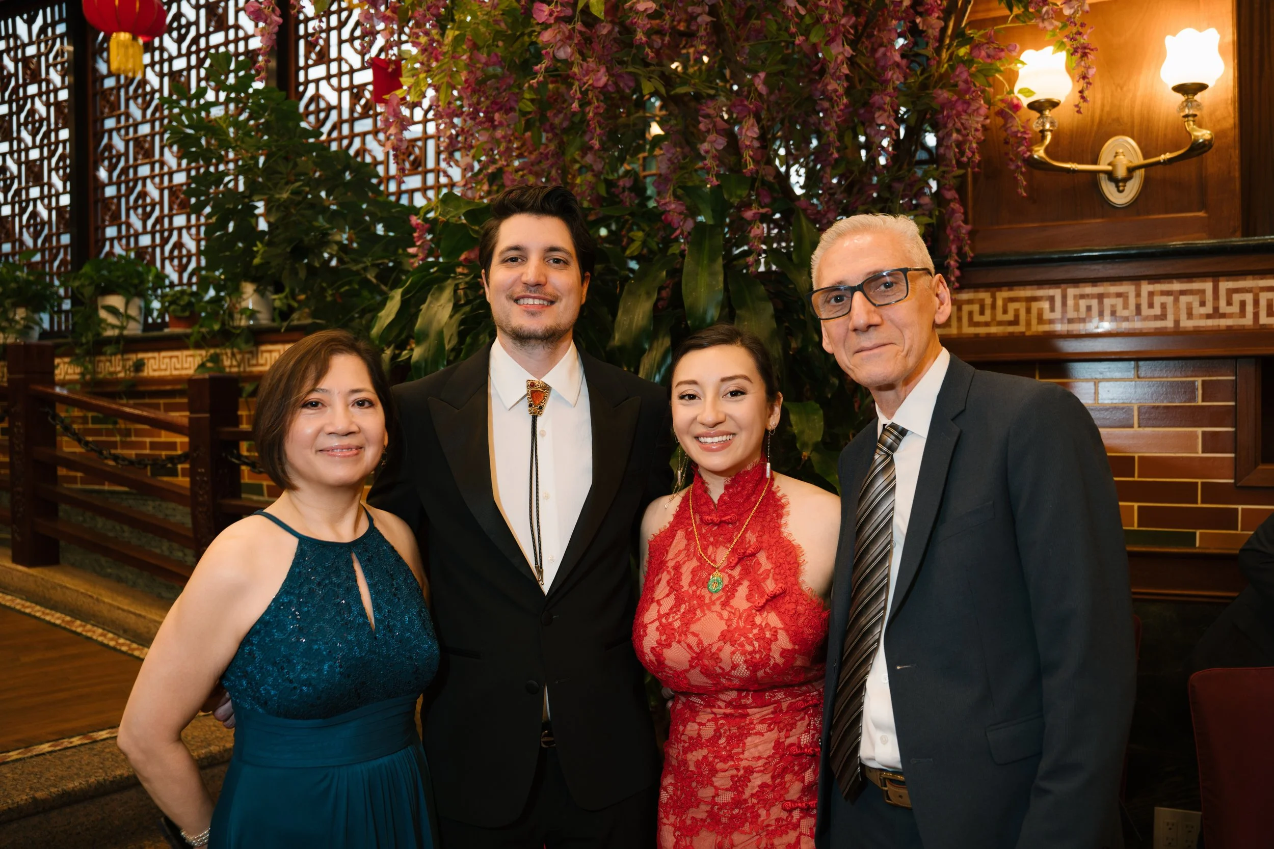 A group of five people dressed in formal attire posing inside a decorated venue with plants and wooden panels.
