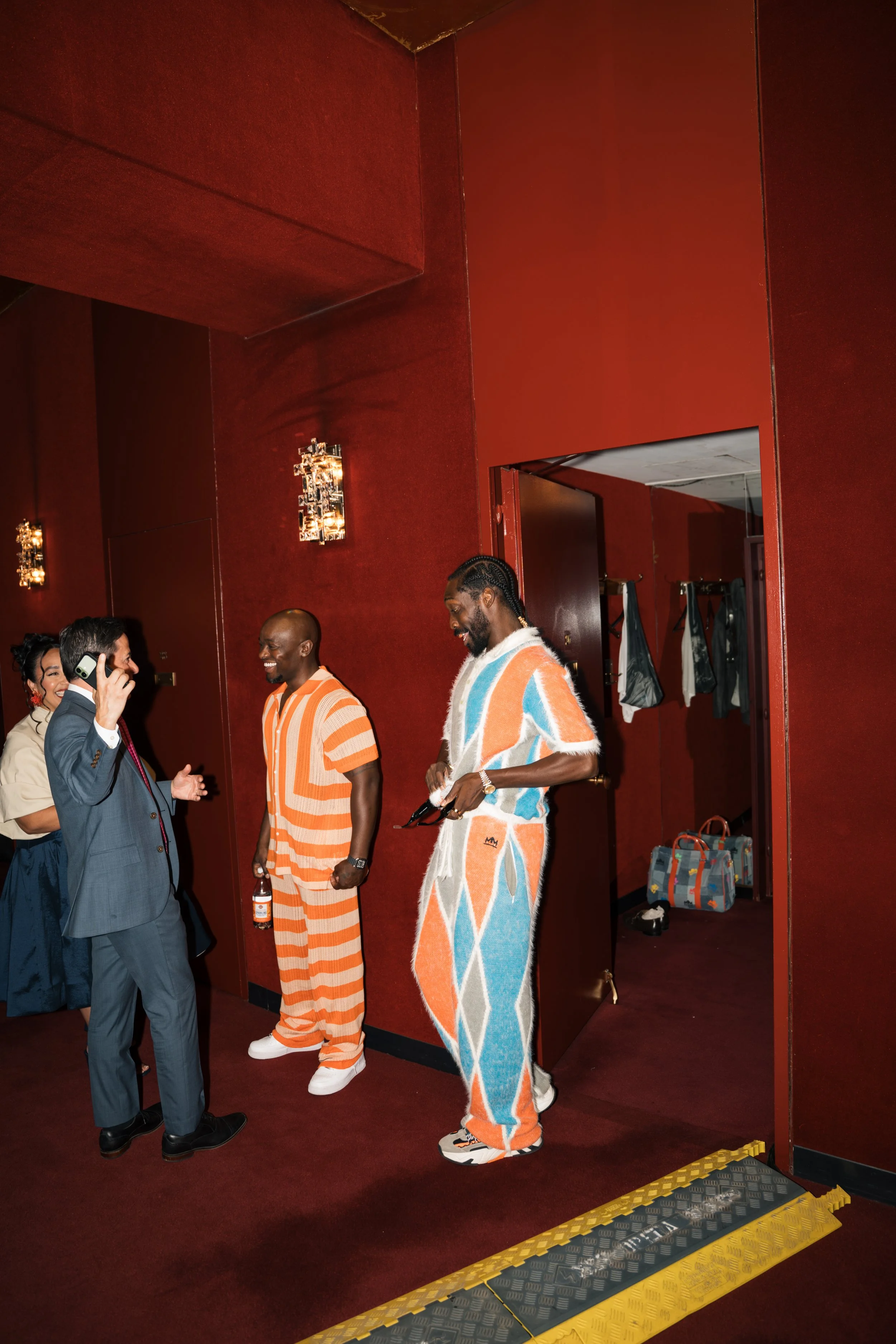 Group of fashionably dressed people talking and laughing in a red-walled room, with a coat rack and bags in the background.