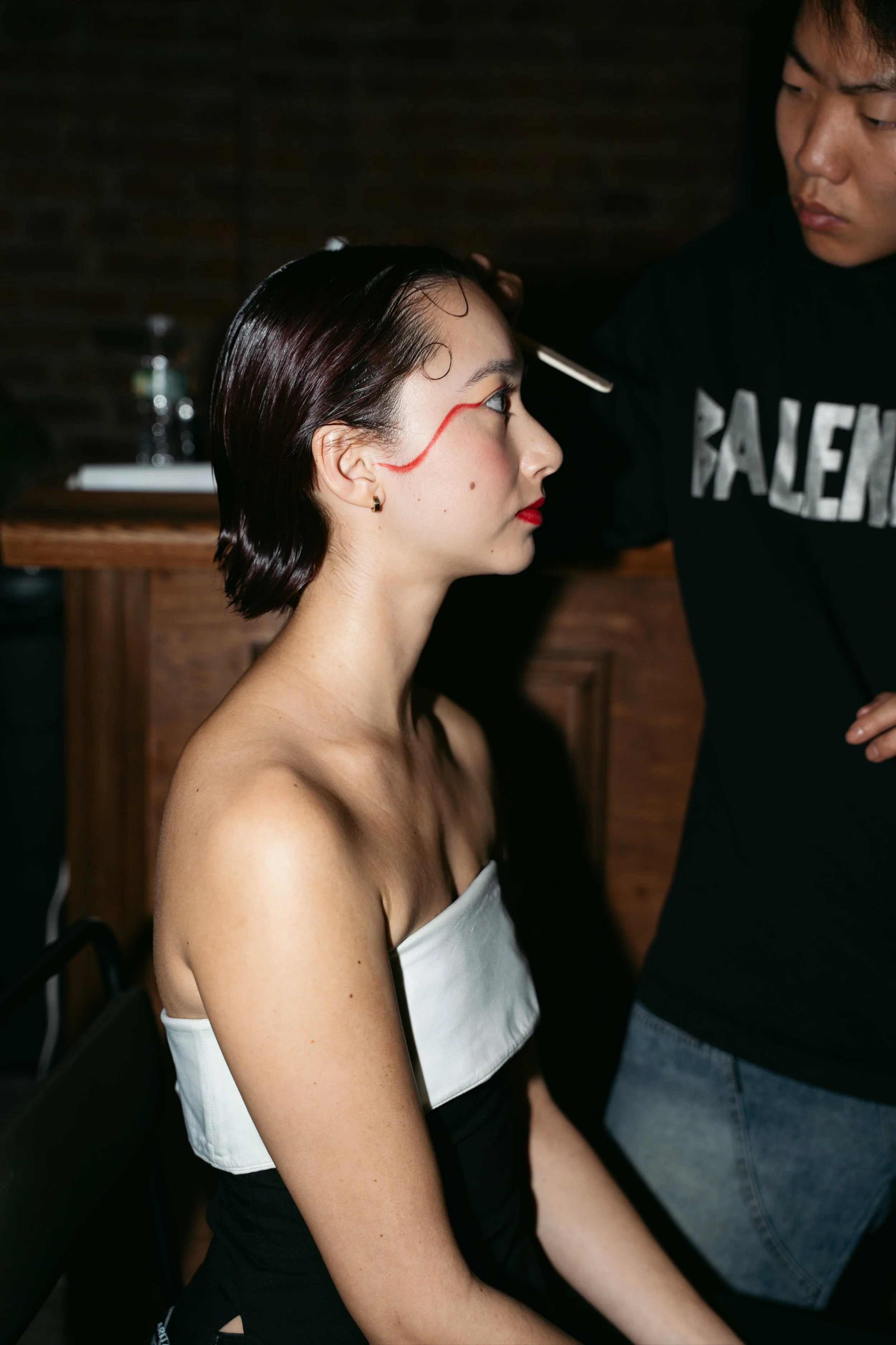 A woman with dark hair and red lipstick is having makeup applied by a person standing next to her. The makeup artist is using a brush on her forehead. The woman is wearing a white strapless top, and the background includes a wooden table and a dark b