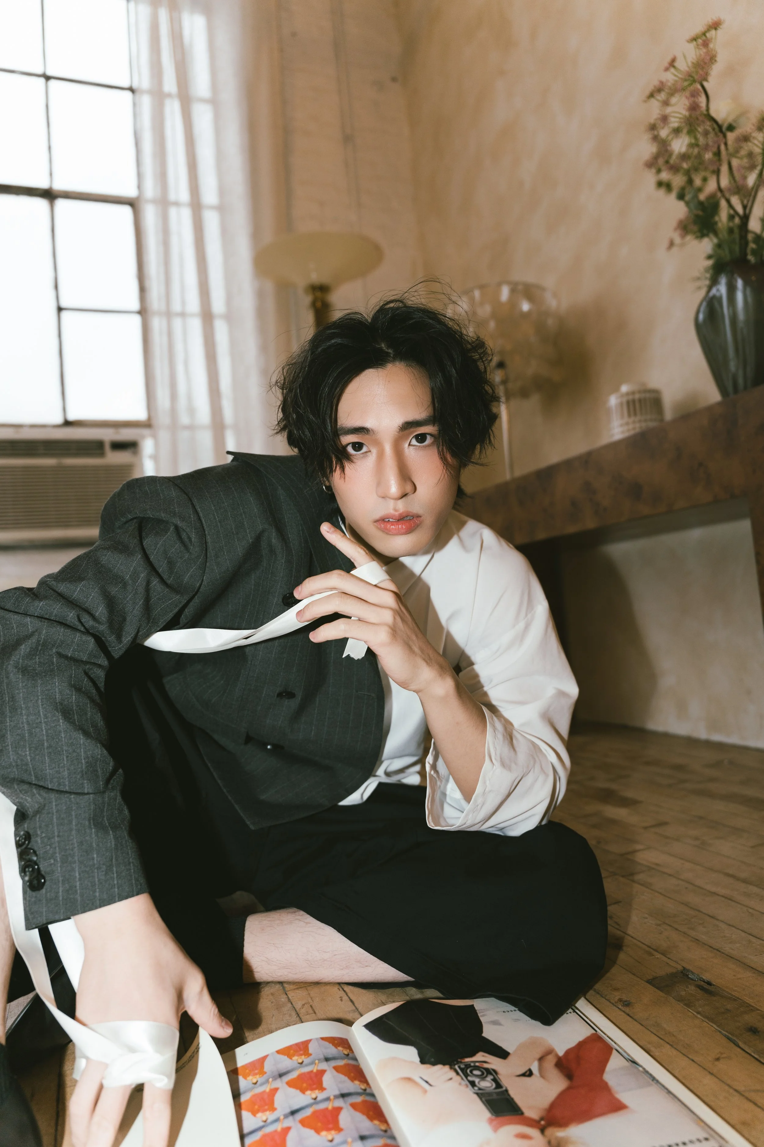 A young man with black wavy hair and fair skin is sitting on the wooden floor, leaning on his left arm. He is dressed in a white shirt and a dark plaid blazer. He is looking directly at the camera with a serious expression. In the background, there i