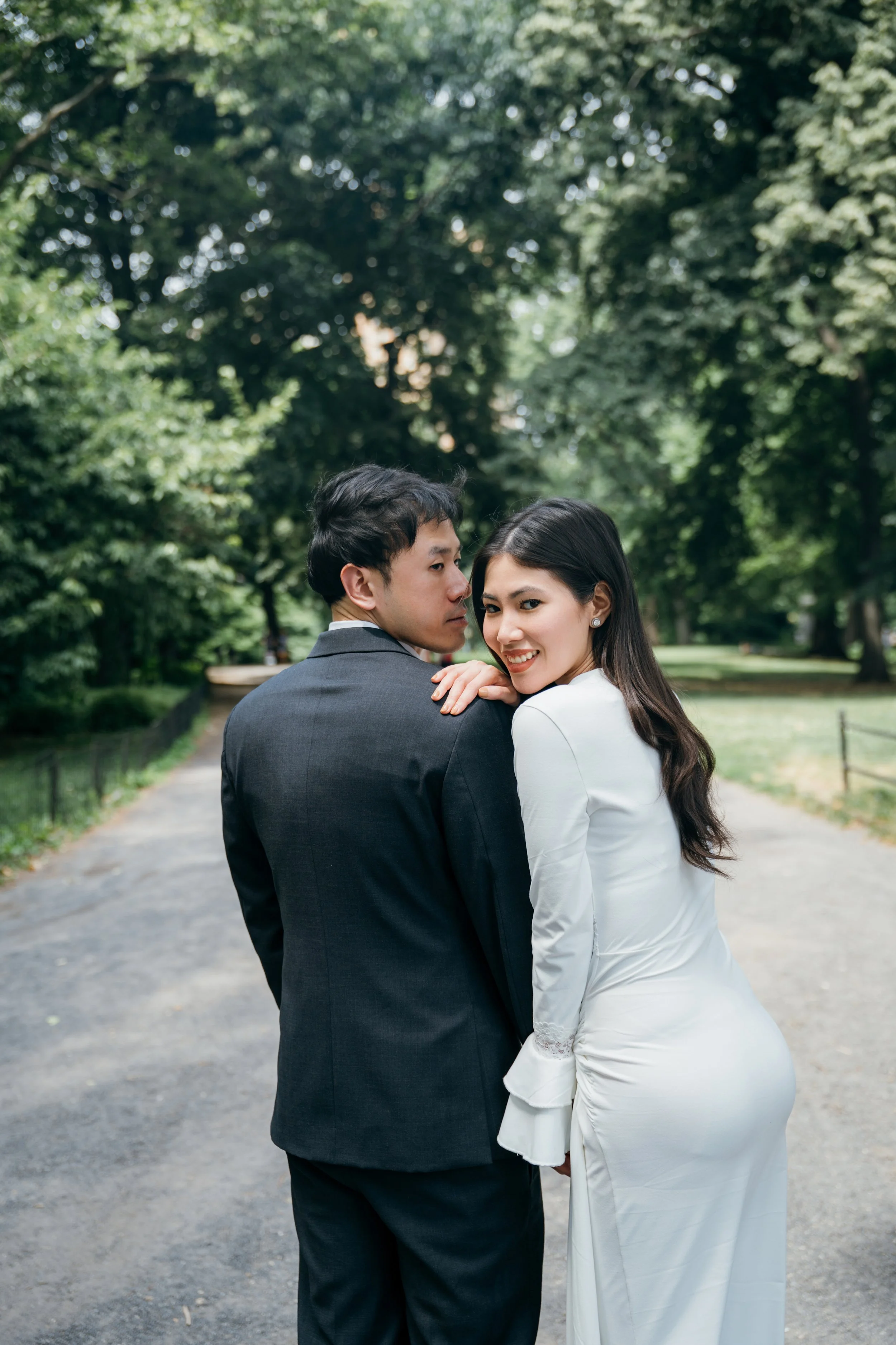 A stylish couple dressed in formal attire, standing close together in a park surrounded by green trees. The woman is smiling at the camera, while the man is looking at her.