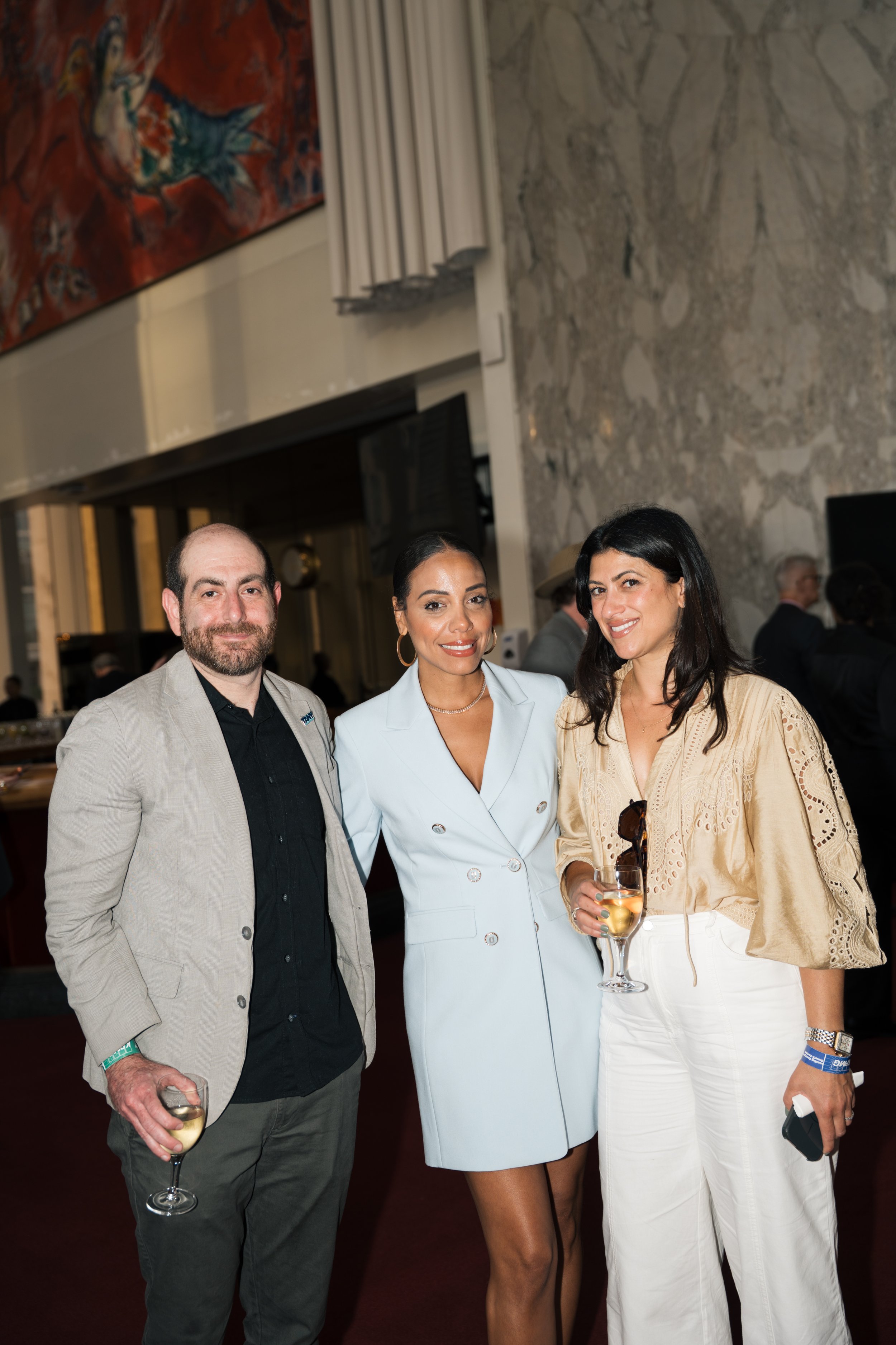 Three people at a social event, two women and one man, holding glasses of wine, smiling and dressed in semi-formal attire, with a textured stone wall and some artwork visible in the background.