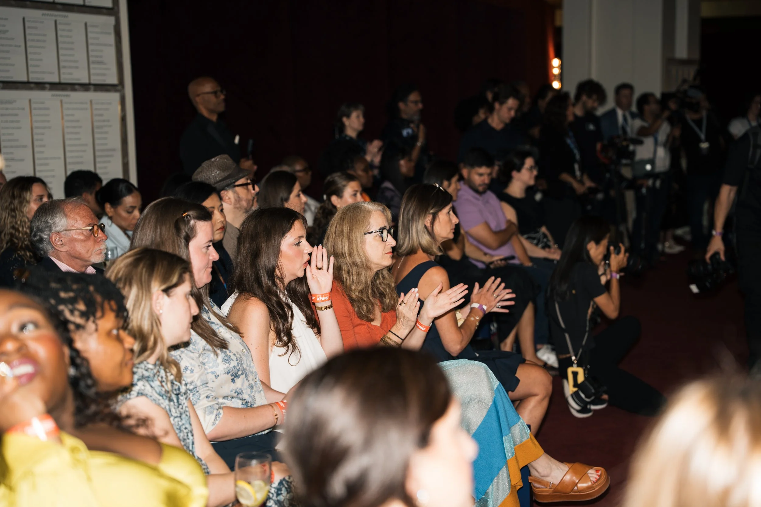Audience seated and watching a presentation at an indoor event, with some audience members clapping.
