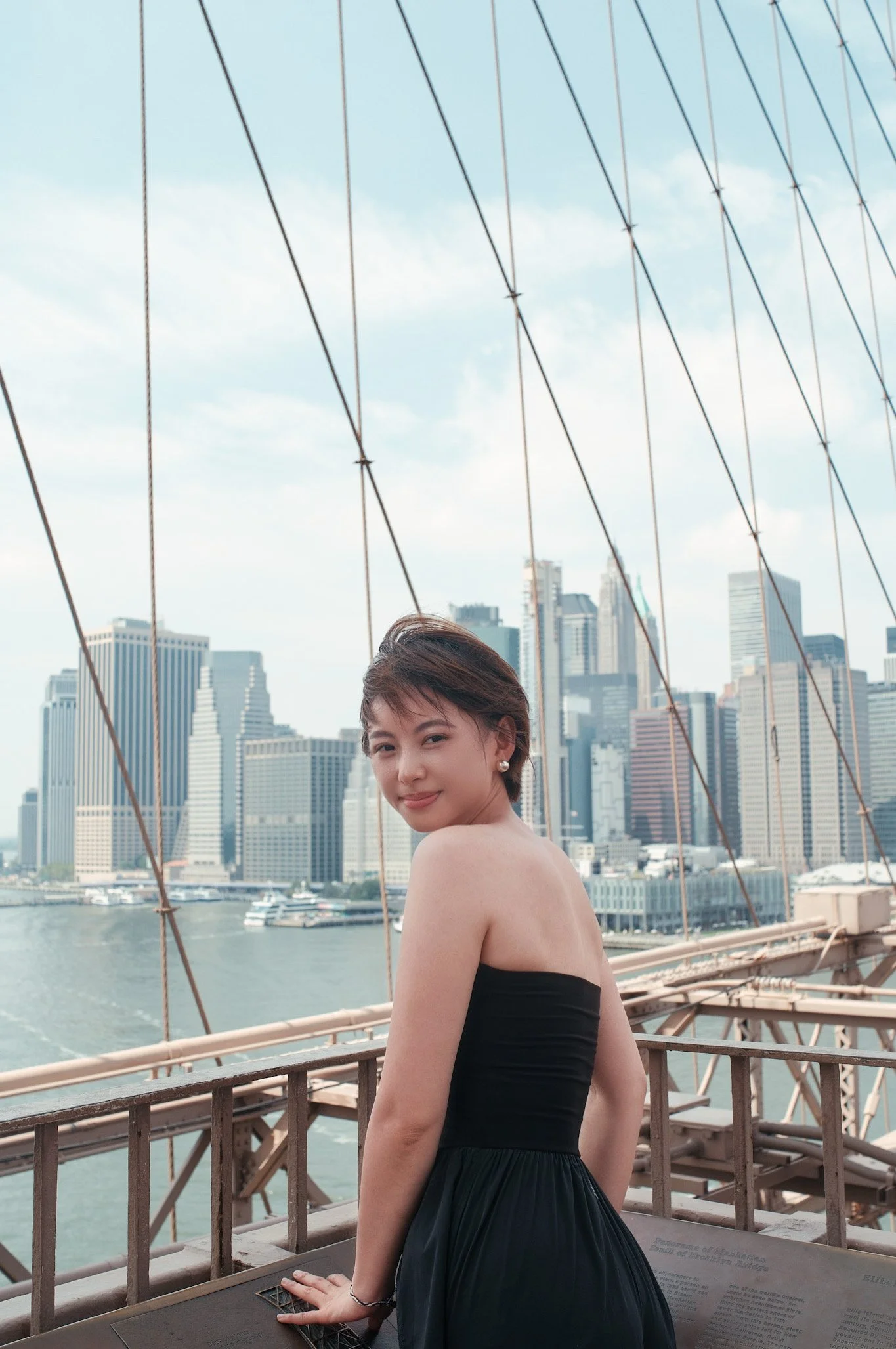 A woman standing on a bridge overlooking a city skyline with tall buildings and a river beneath, with wires and part of the bridge structure in view.