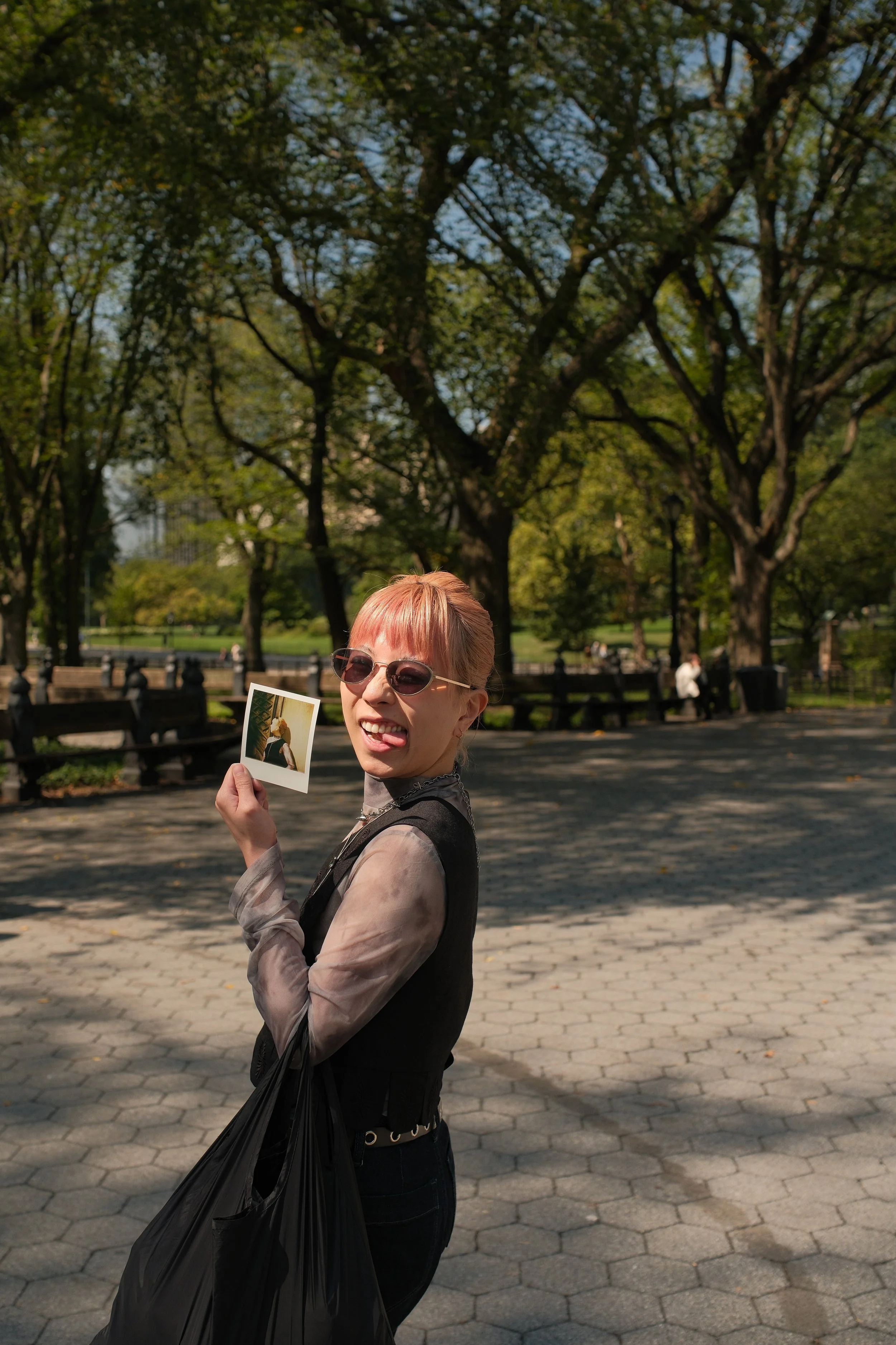 A woman with short hair, wearing sunglasses, smiling and sticking out her tongue, holding a photo while standing outdoors in a park with trees and benches in the background.