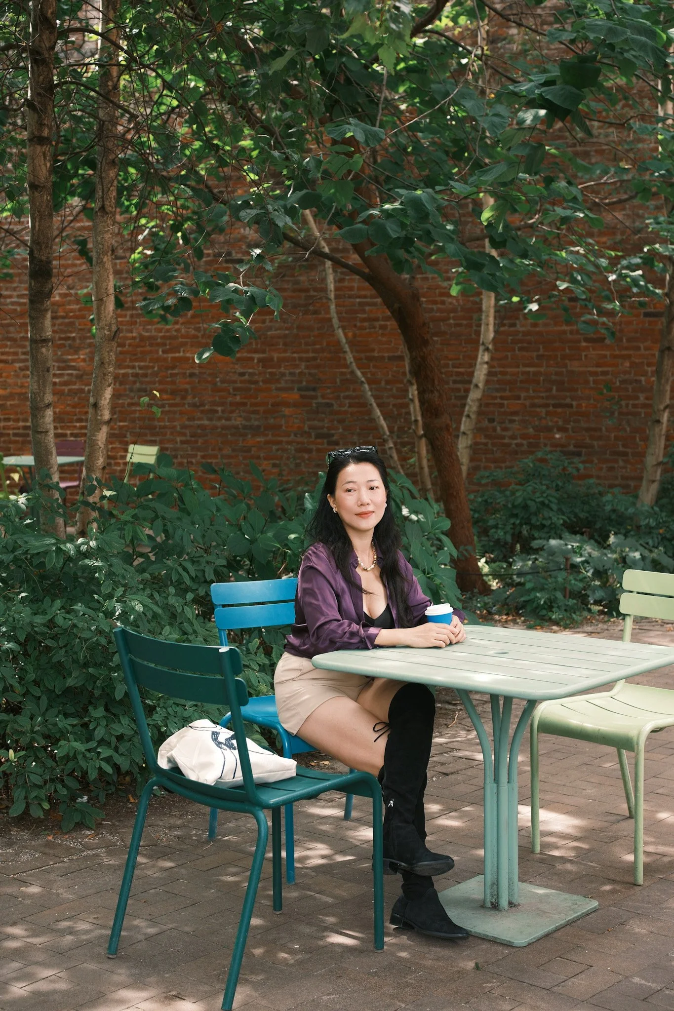 A woman sitting at an outdoor table in a park, holding a blue coffee cup, with greenery and trees around her and a brick wall in the background.
