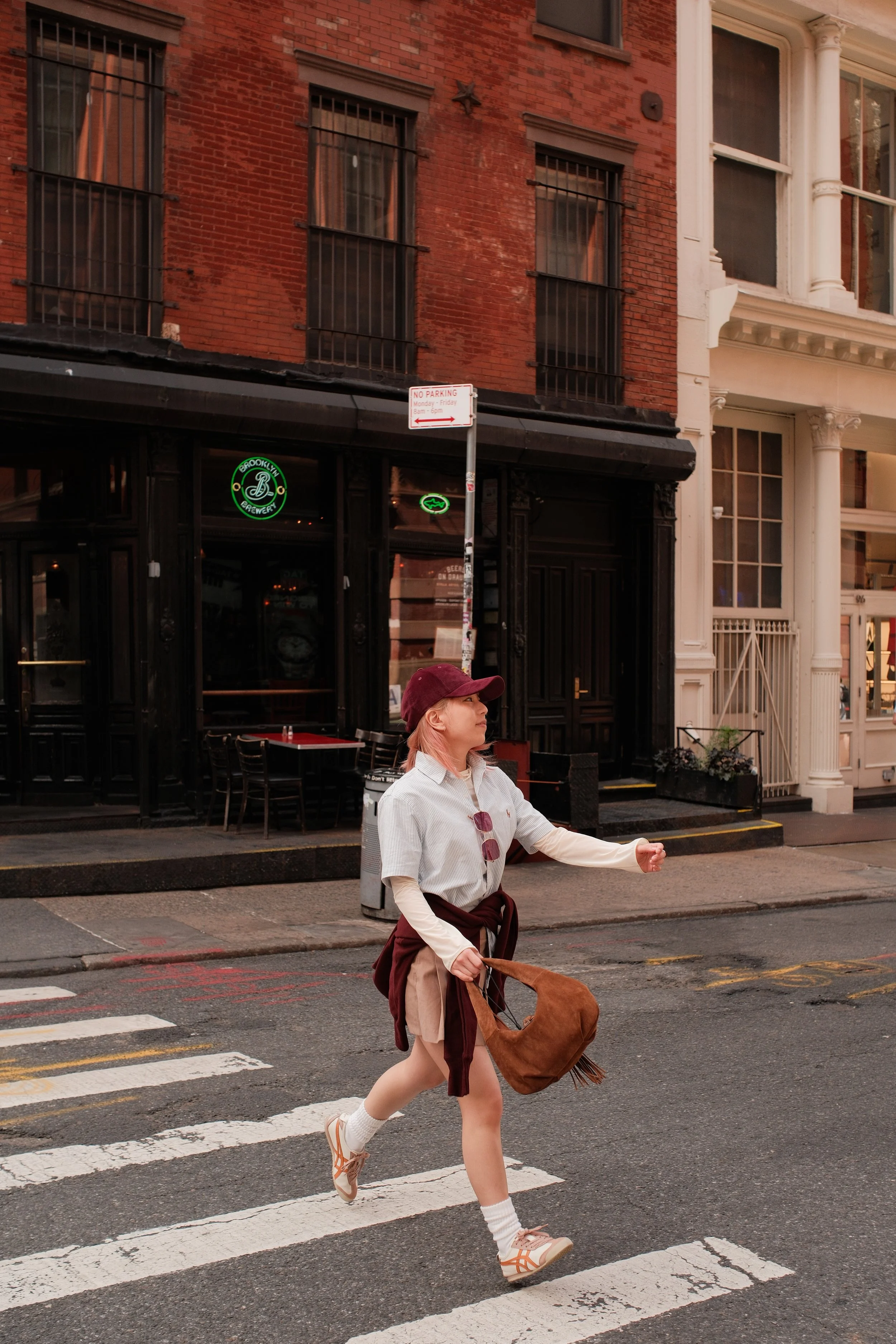A woman crossing the street on a crosswalk in an urban setting, carrying a brown bag and wearing a burgundy hat, beige shorts, white socks, and sneakers, with brick and white buildings in the background.