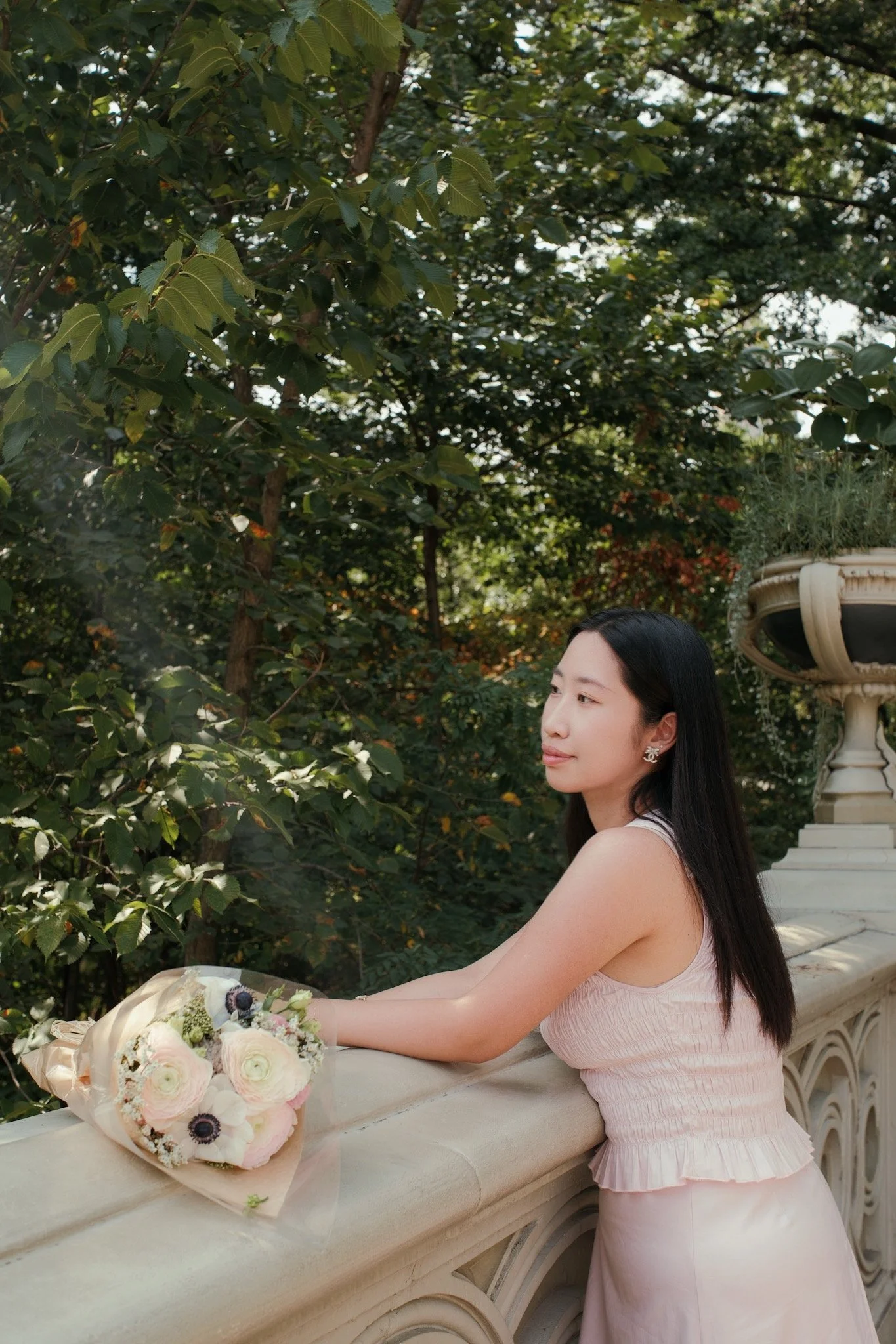 A woman with long dark hair and earrings wearing a light pink dress with ruffled waistline, standing outdoors near a stone balustrade with a bouquet of flowers resting on it, surrounded by green trees and foliage.