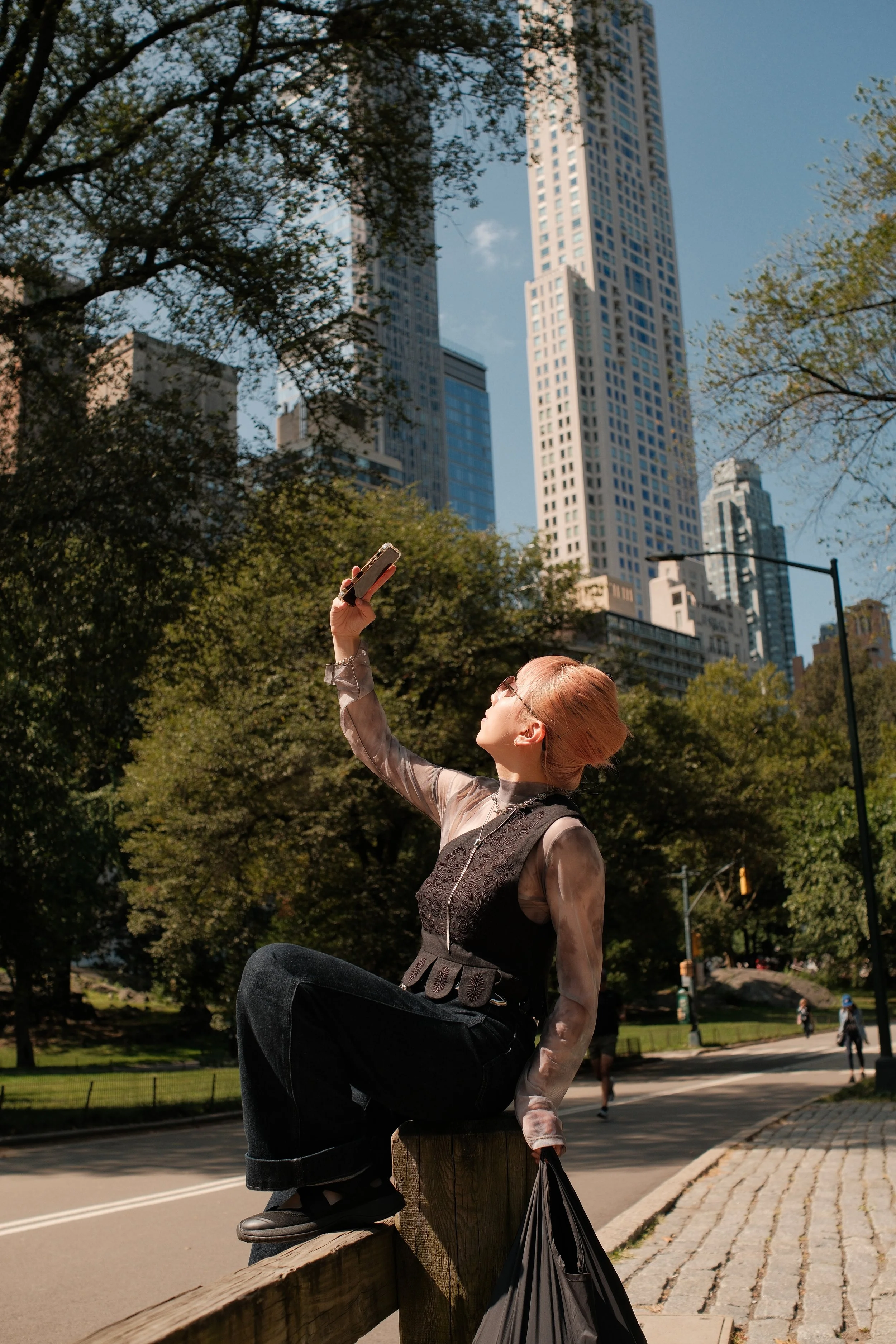 A woman with reddish hair and glasses sitting on a fence in a park, taking a selfie with tall skyscrapers and trees in the background.