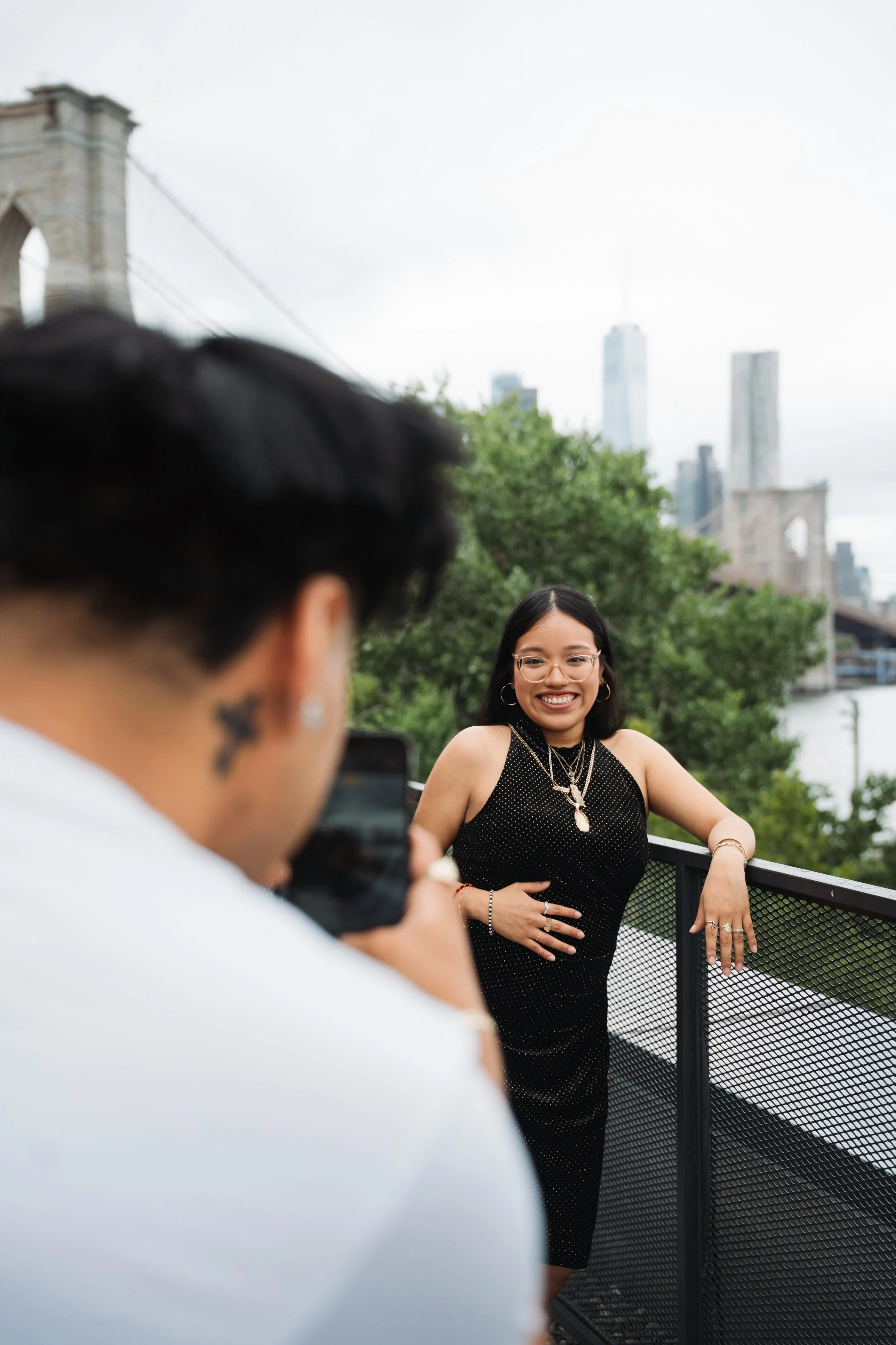 A woman in a black dress with jewelry posing on a balcony outdoors while a person in the foreground takes her photo. A city skyline with tall buildings and Brooklyn Bridge is in the background.