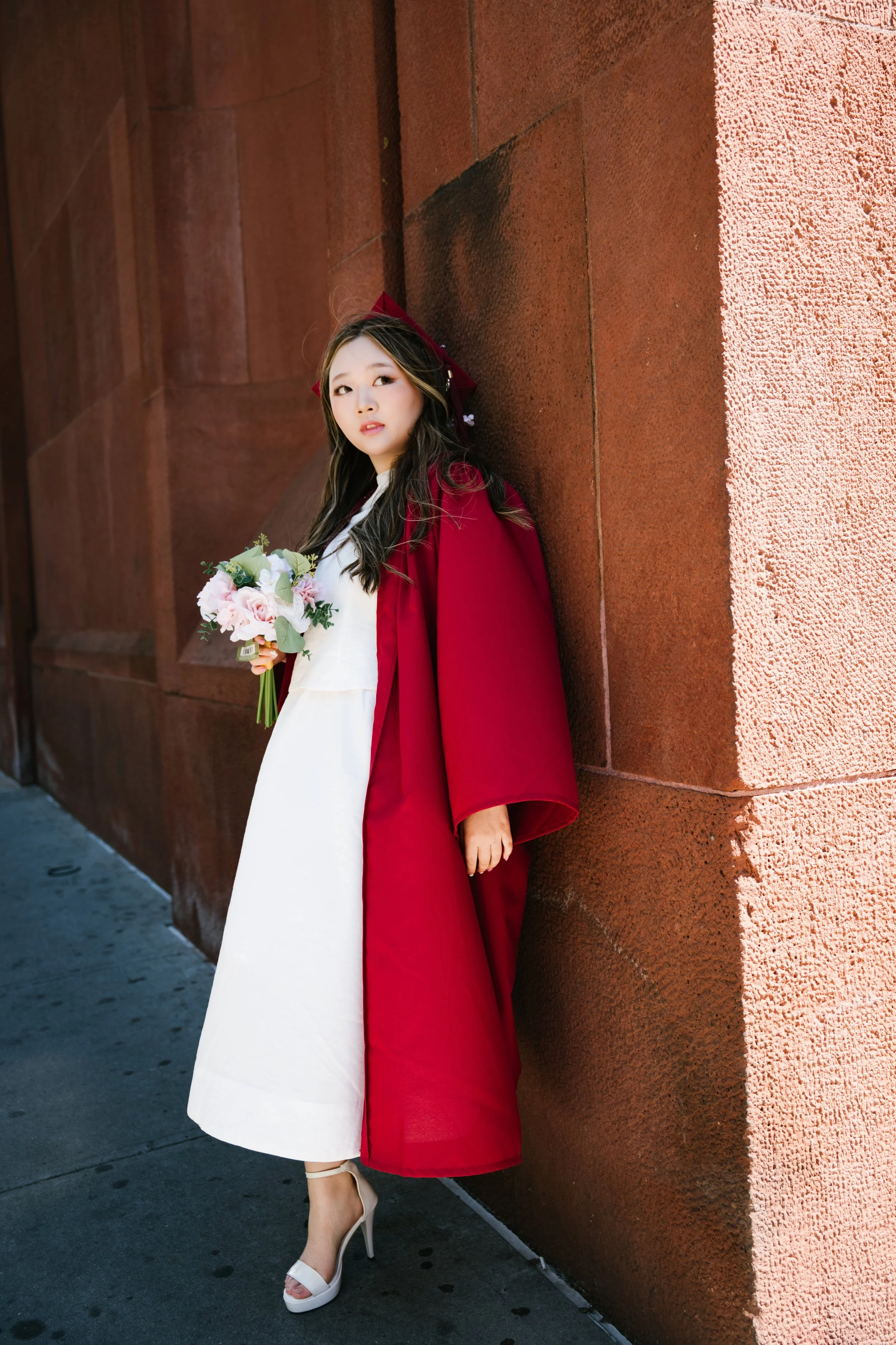 Young woman in a white dress, red cape, and white high heels standing against a red brick wall, holding a bouquet of pink and white flowers.